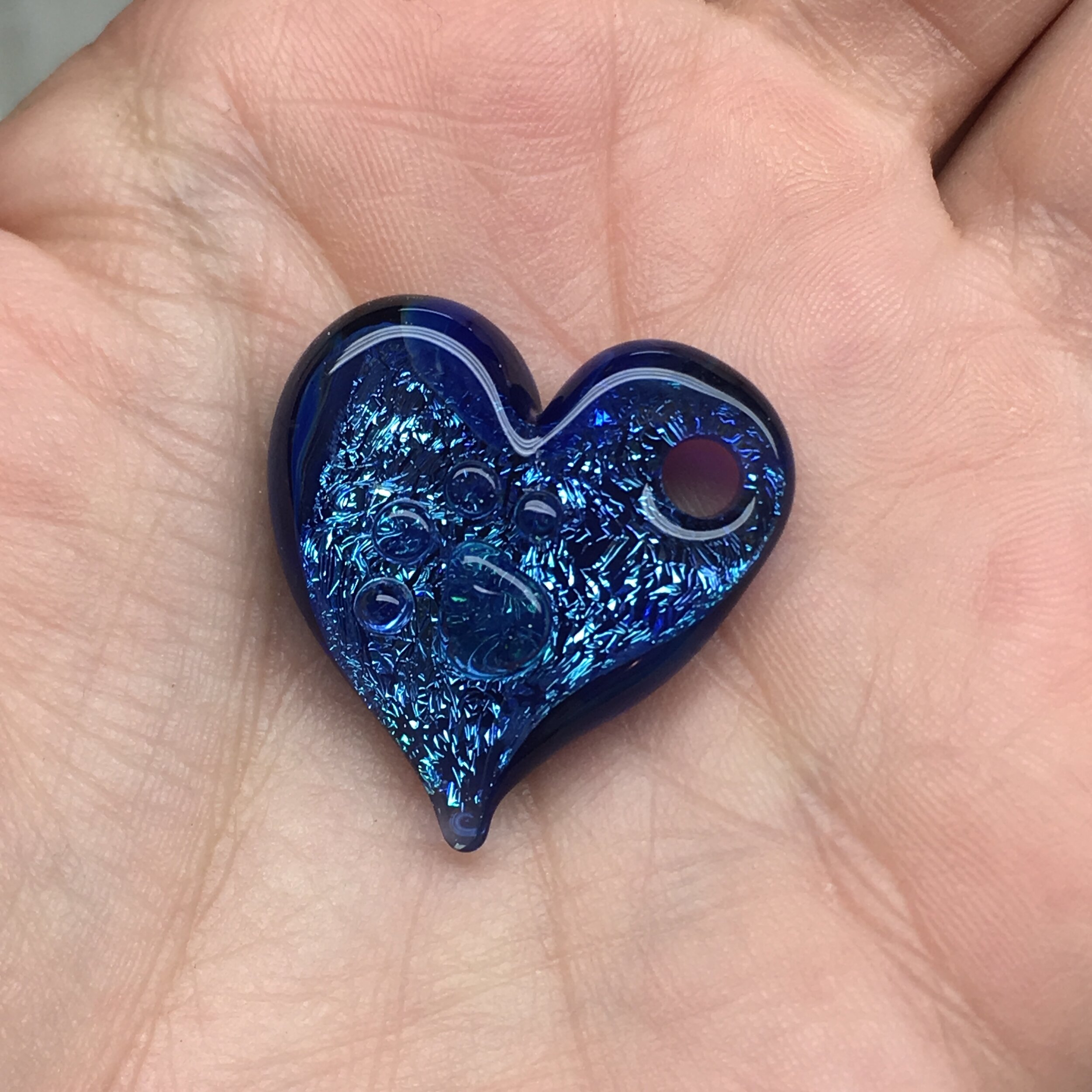 A small, blue, glass heart with bubble and crackle effects, held in a person's hand.
