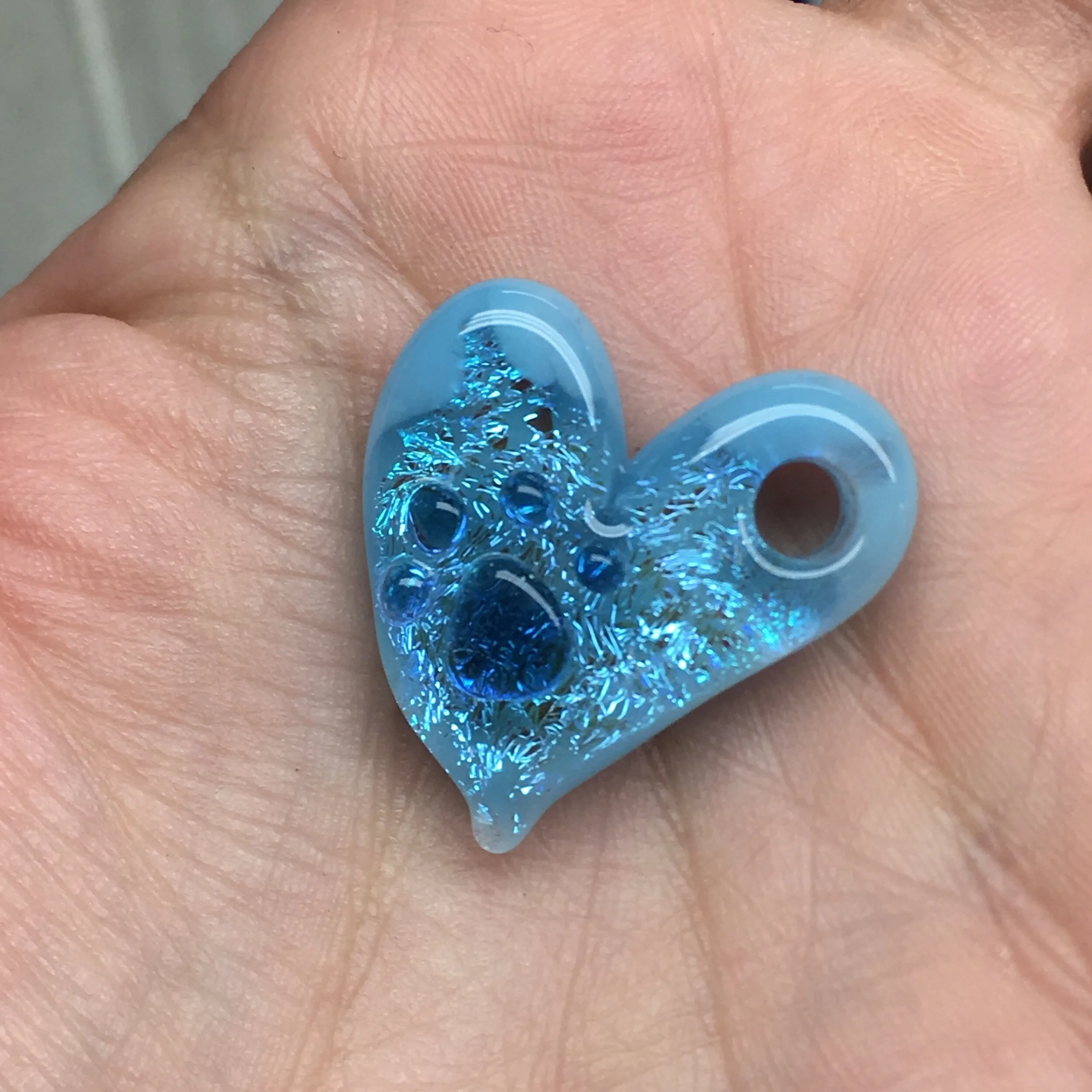Close-up of a blue, heart-shaped, glittery bead with holes, held in a person's palm.