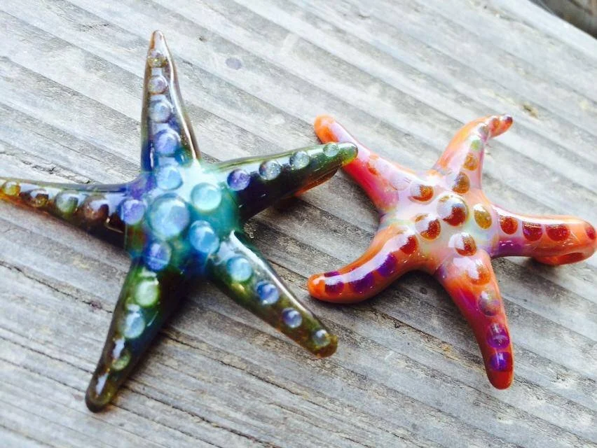 Two colorful glass starfish decorations on a wooden surface, one blue-green and the other orange with purple spots.