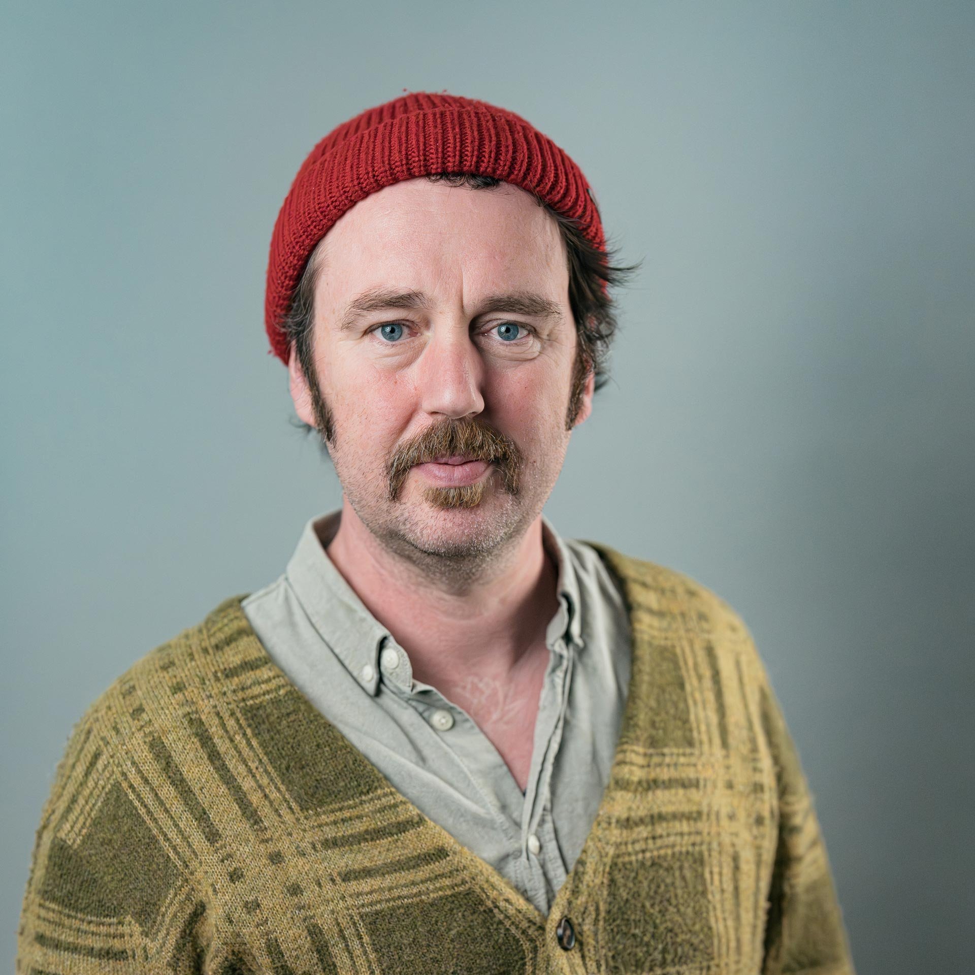 Nahaufnahme eines Mannes mit hellblauen Augen, braunem Bart und längeren braunen Haaren, trägt einen roten Wollmütze und einen grünen Pullover, vor einem grauen Hintergrund.