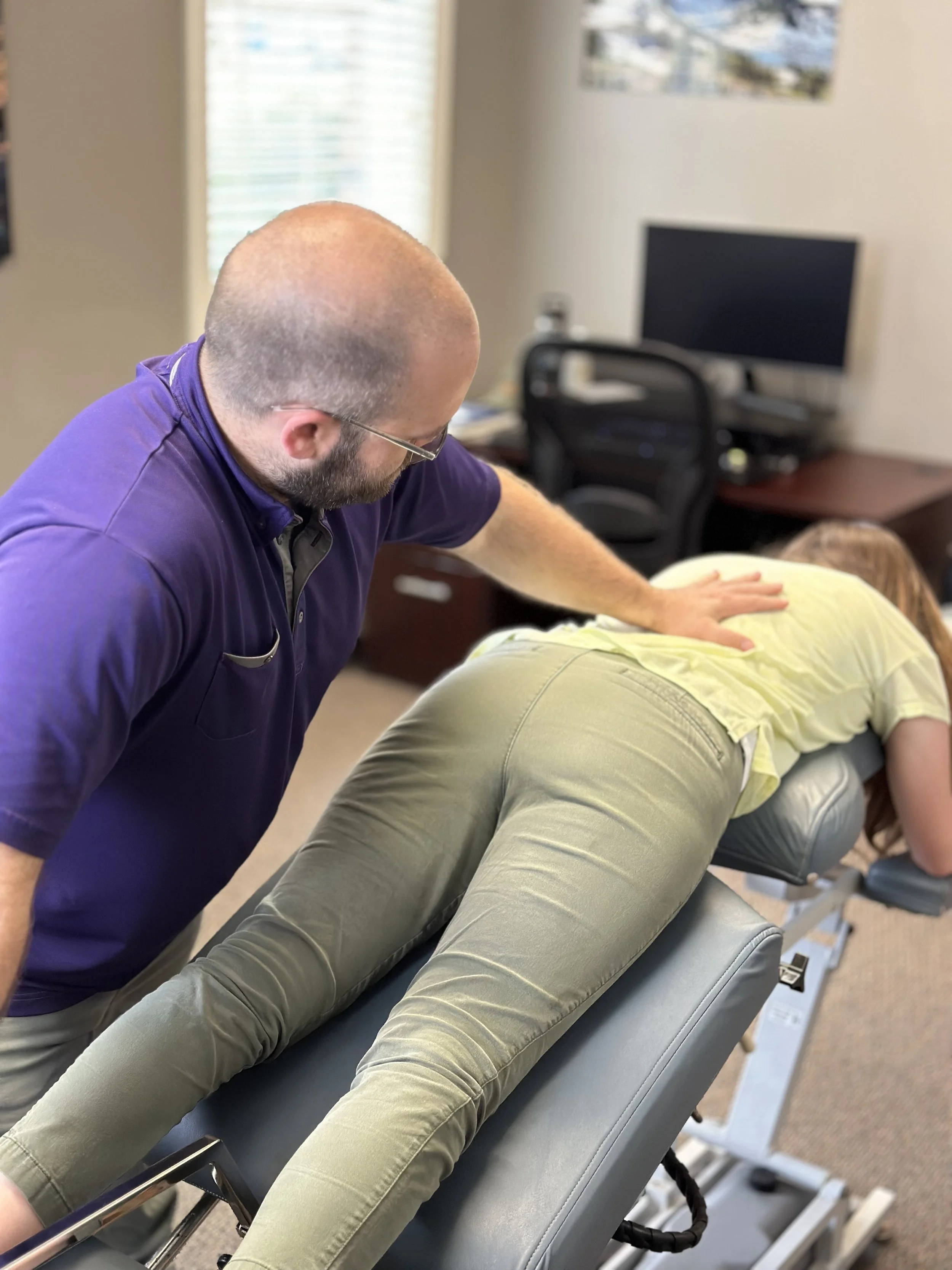A chiropractor adjusts a patient lying face down on an examination table in an office setting.