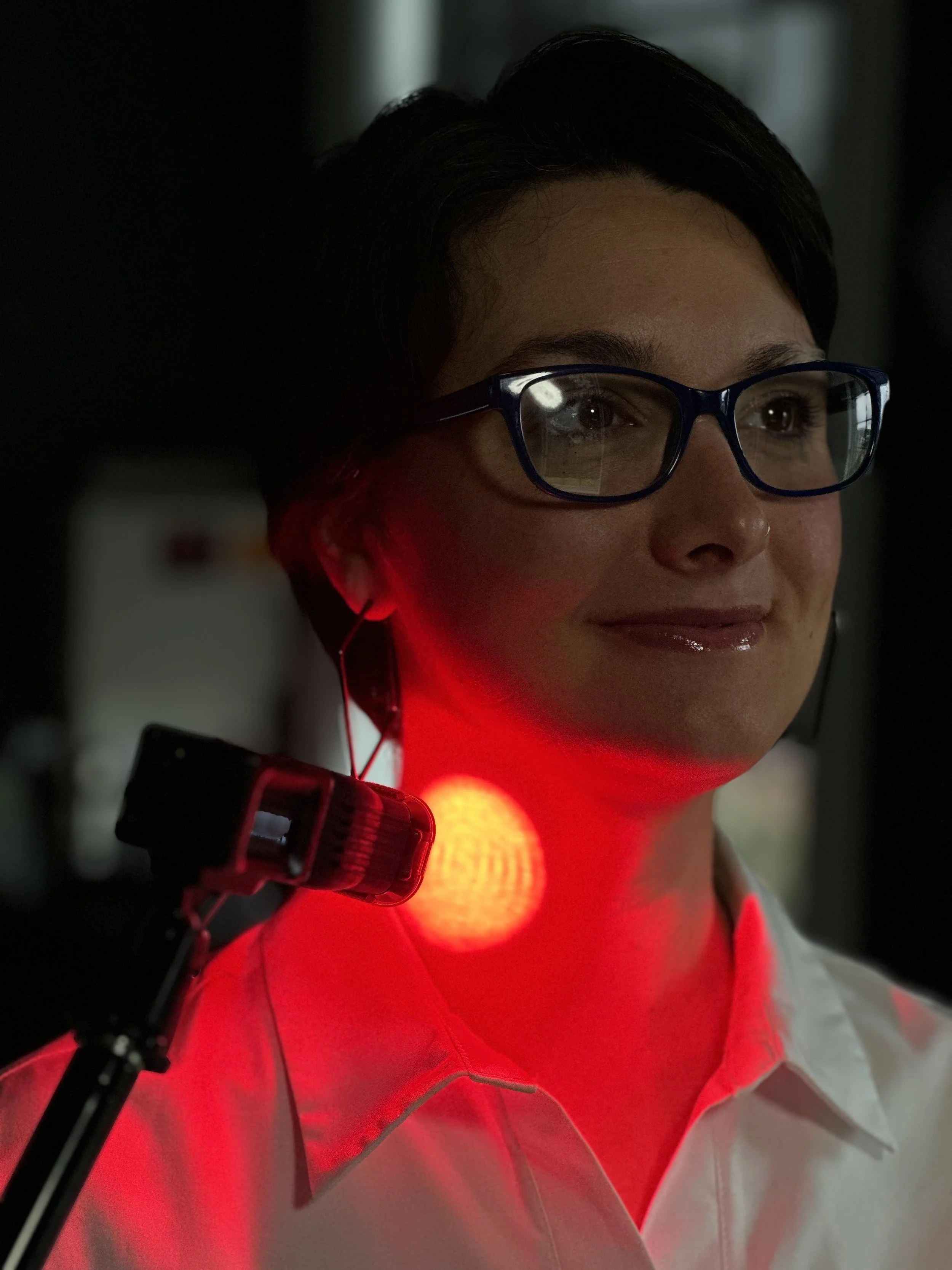 A woman with short dark hair and glasses wearing a white shirt, standing in a dark environment, illuminated by a red light on her neck and shoulder.