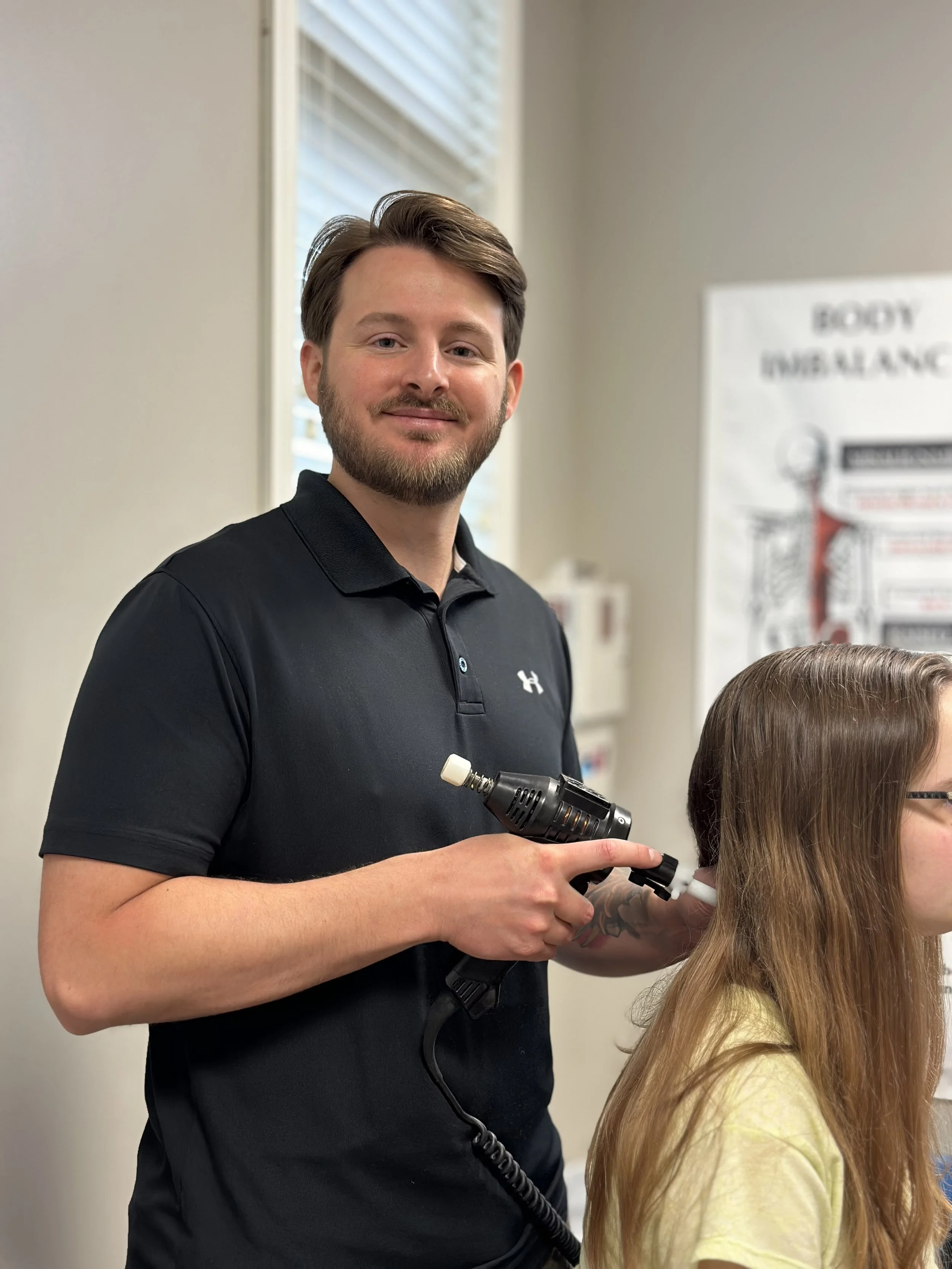 A man with a beard and brown hair, wearing a black polo shirt, is holding a heat gun near a girl's hair in a classroom.