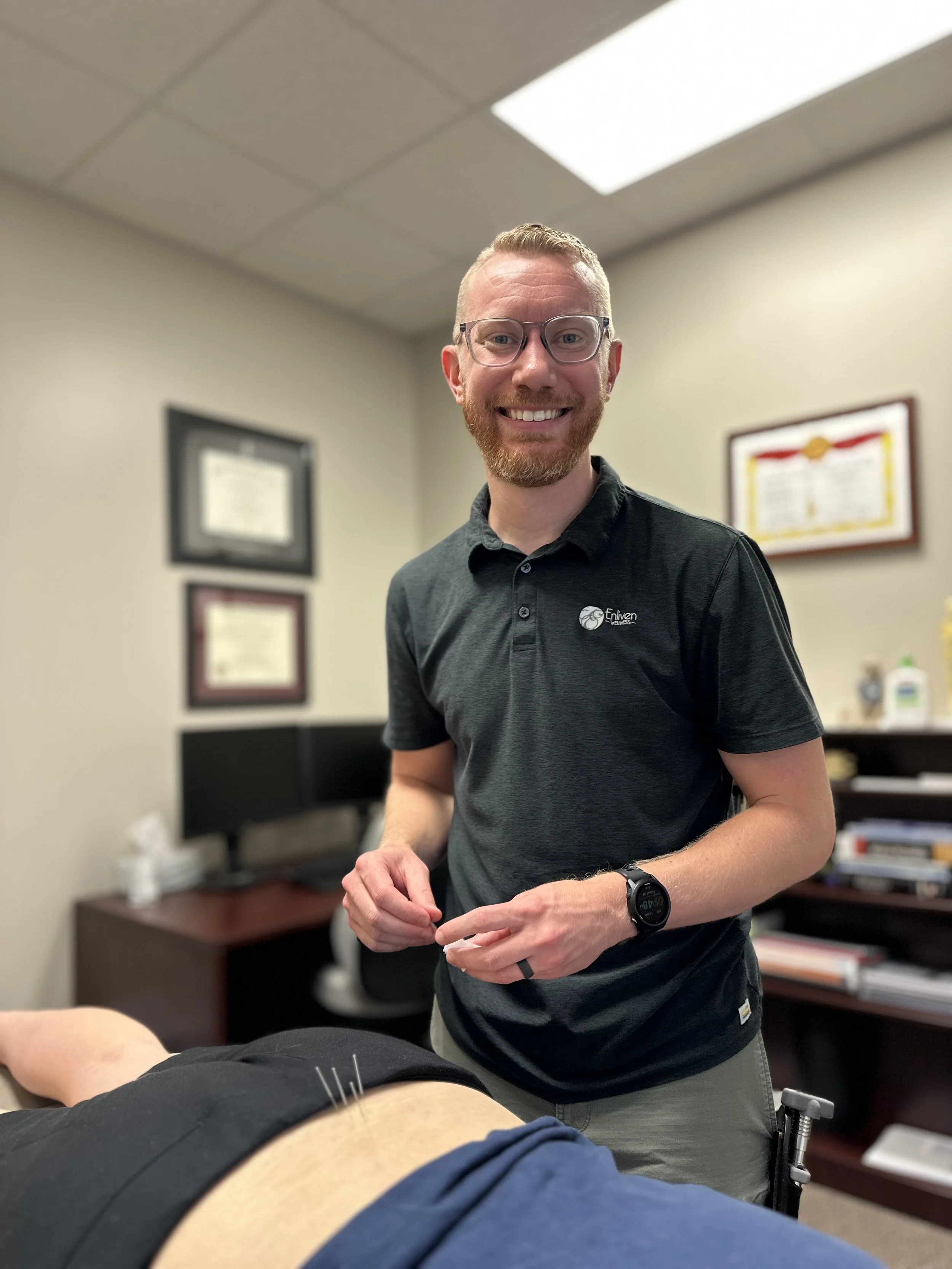 A smiling man with glasses and a beard, wearing a black polo shirt, standing in a medical office near a patient undergoing acupuncture treatment.