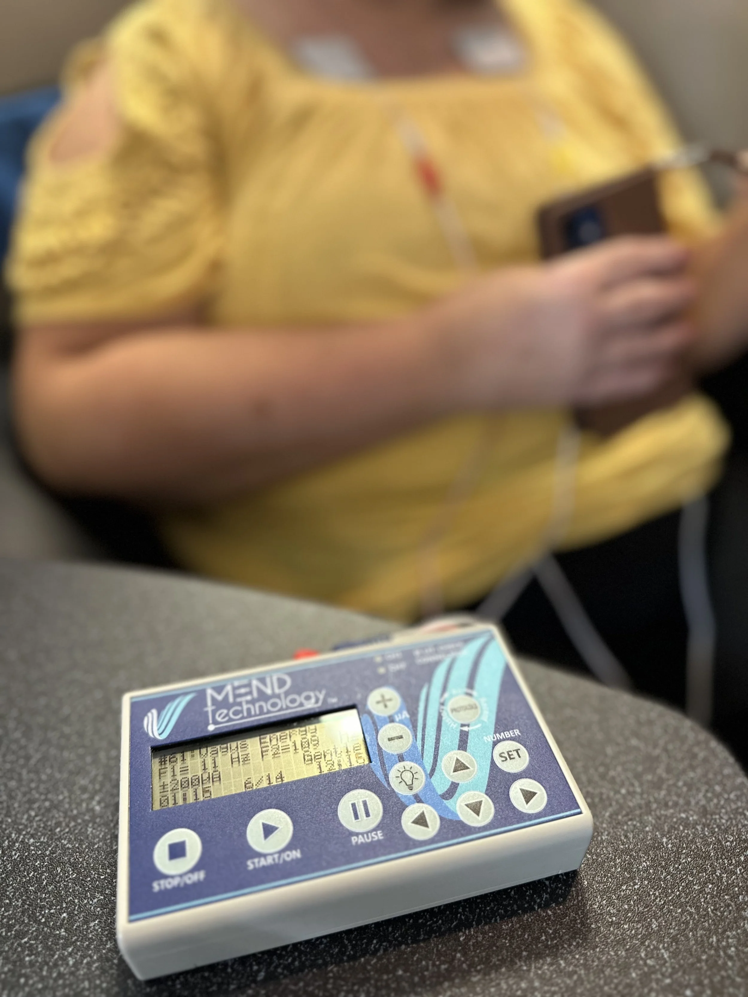 A close-up of a medical device labeled 'MEND technology' on a black surface, with a person in a yellow shirt using a smartphone in the background.
