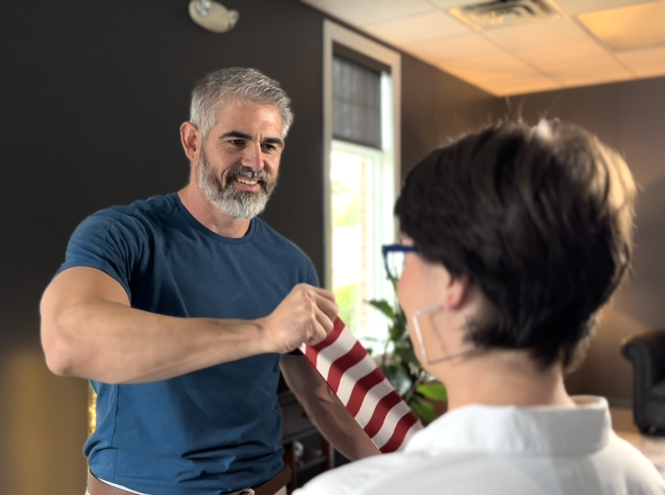 A man with gray hair and a beard is smiling as he hands a gift wrapped in red and white striped paper to another person with short dark hair and glasses, inside a room with dark walls and a window.