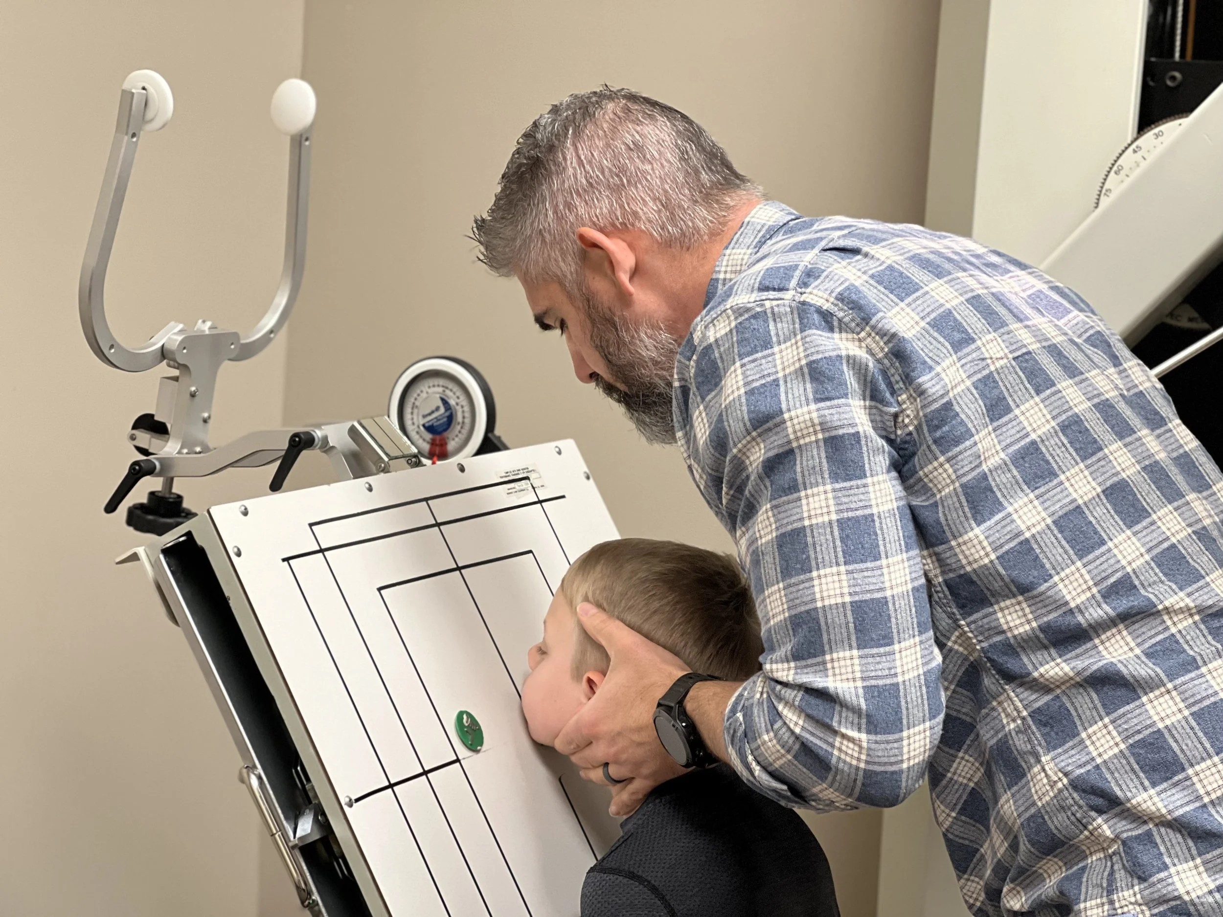A man with gray hair and beard conducting an ear, nose, and throat test on a young boy at a medical clinic.