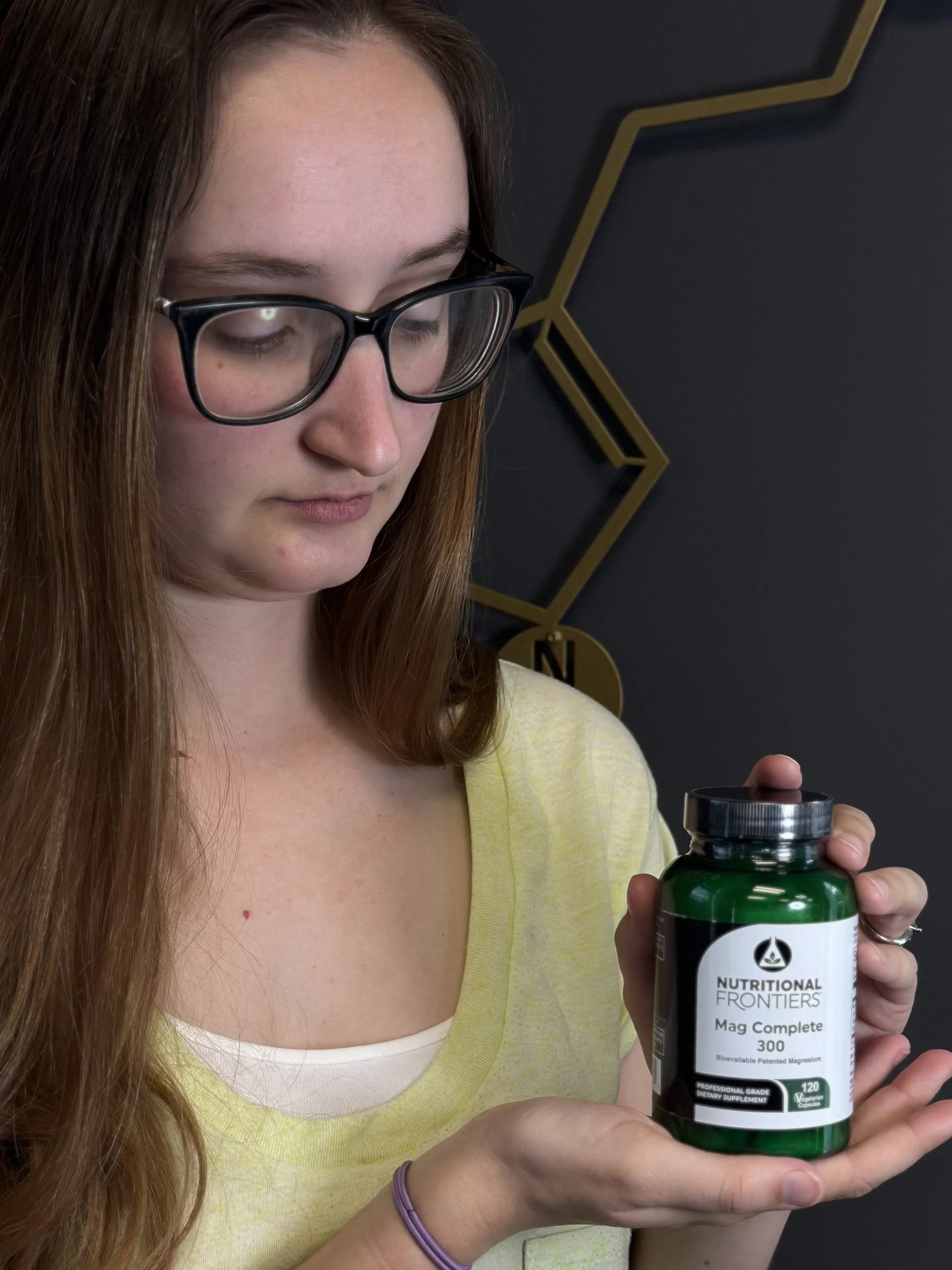 A young woman with long brown hair, wearing glasses and a yellow shirt, holding a bottle of Nutritional Frontiers Mag Complete 300 dietary supplement.