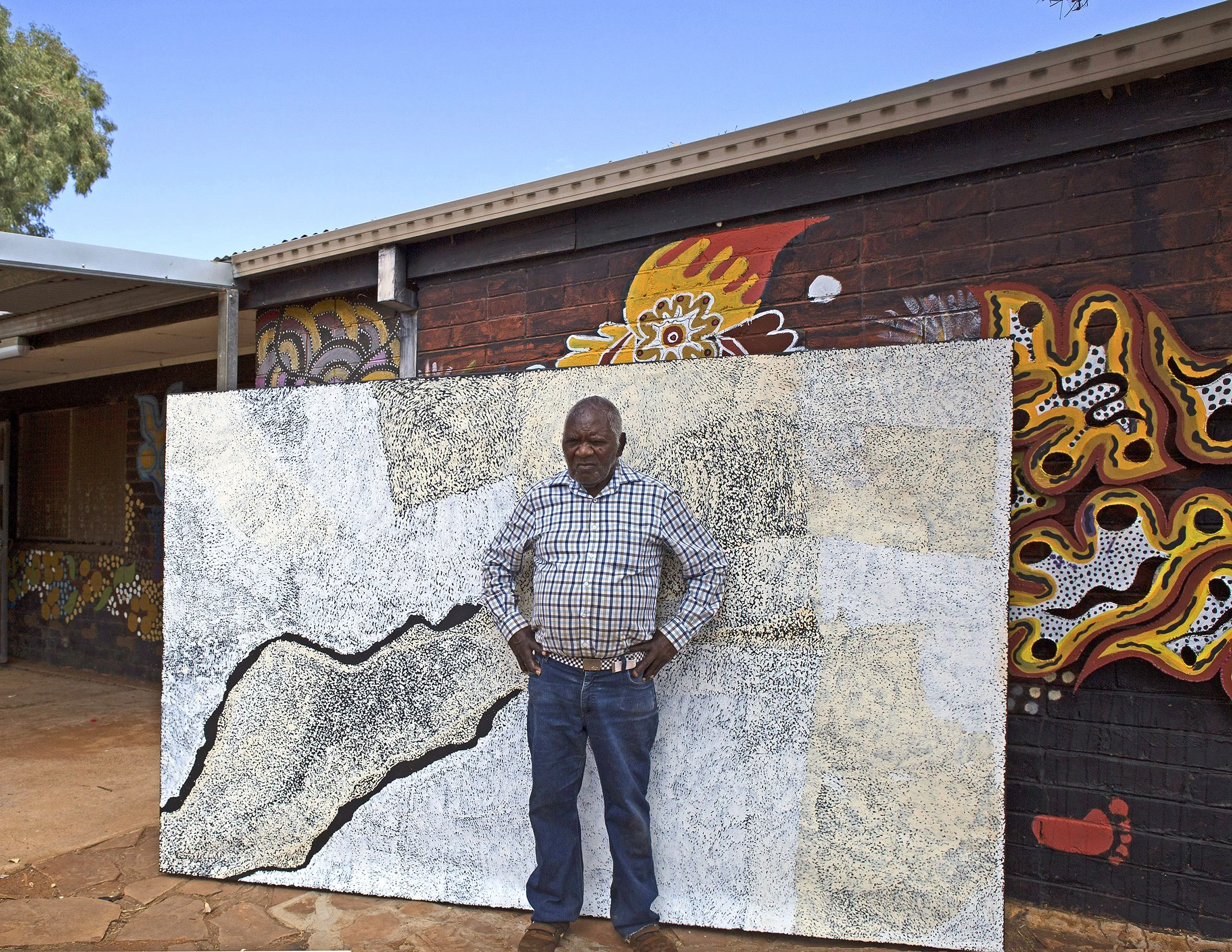 Pepai Jangala Carroll posant avec son œuvre magistrale, juste devant le mur du centre d'art d'Ernabella dans le APY land. © Photo Ernabella Arts with the courtesy of the artist.