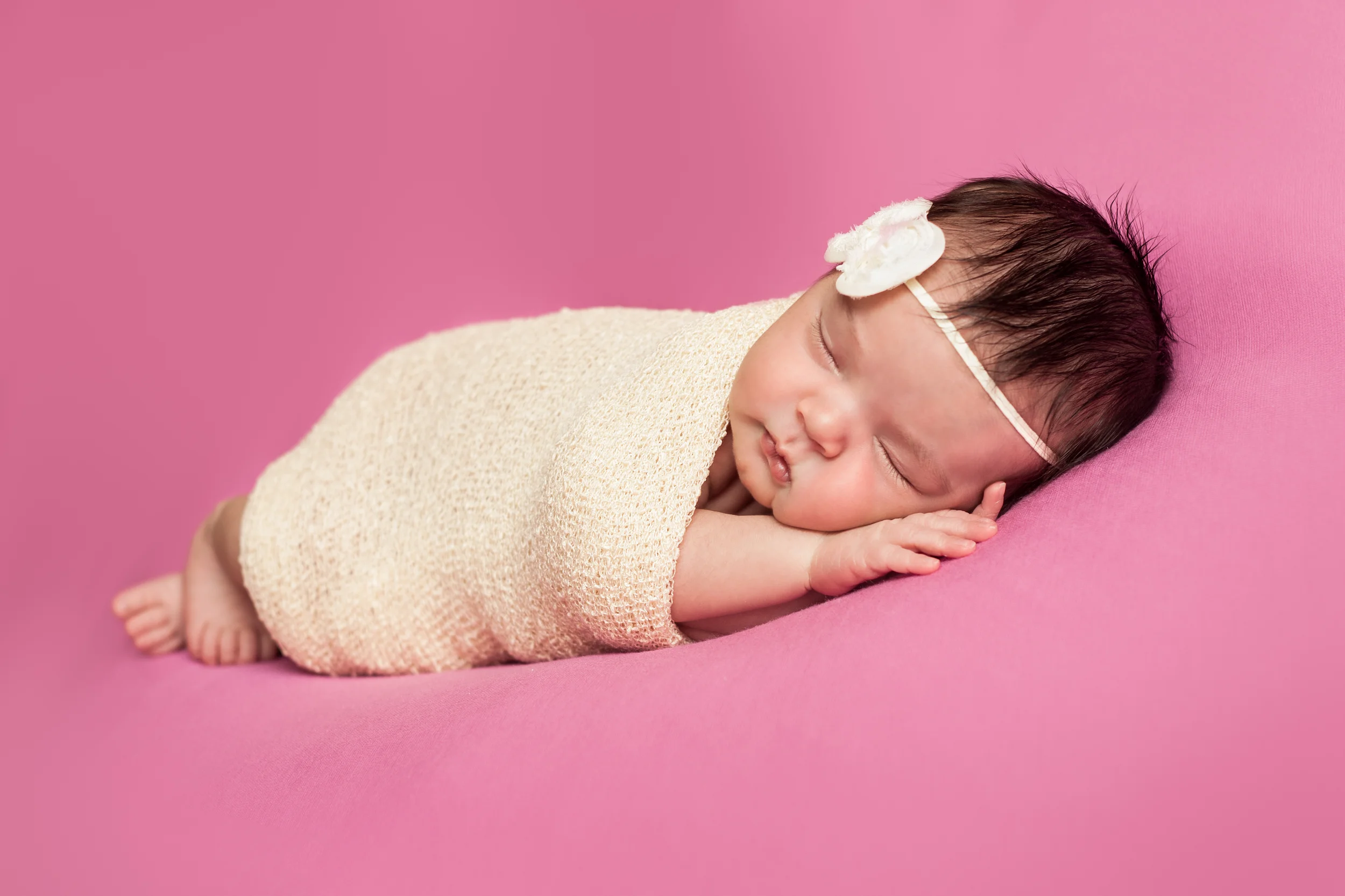 Newborn baby girl wrapped in creme stretch wrap with creme bow headband on pink background, Westchester County, NY Newborn Photographer Amanda Noelle Photography.