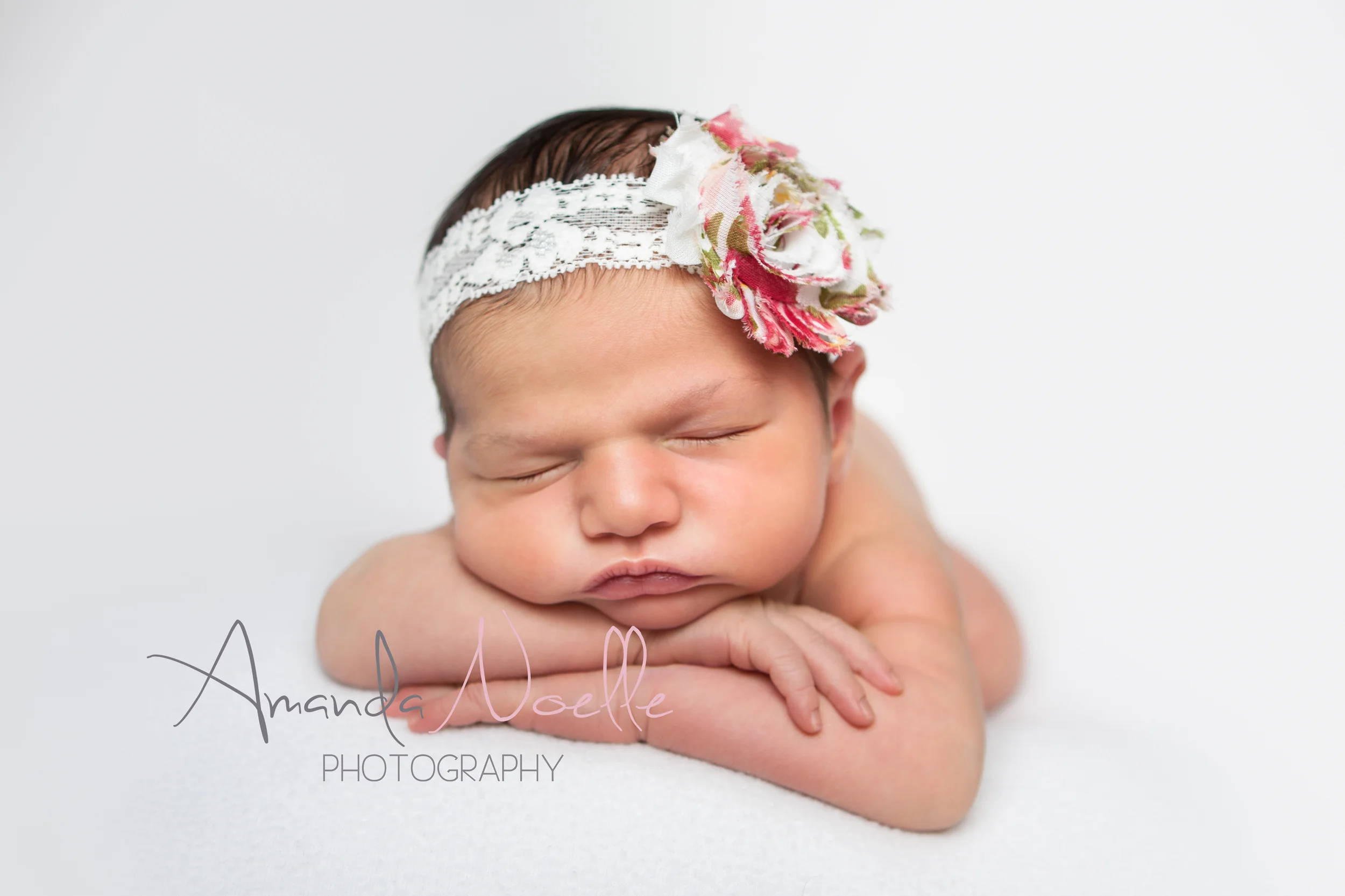 Newborn baby girl, white background fabric, white lace pink shabby chic flower headband, sleeping pose chin on hands on beanbag, by Westchester, New York Newborn Photographer, Amanda Noelle Photography