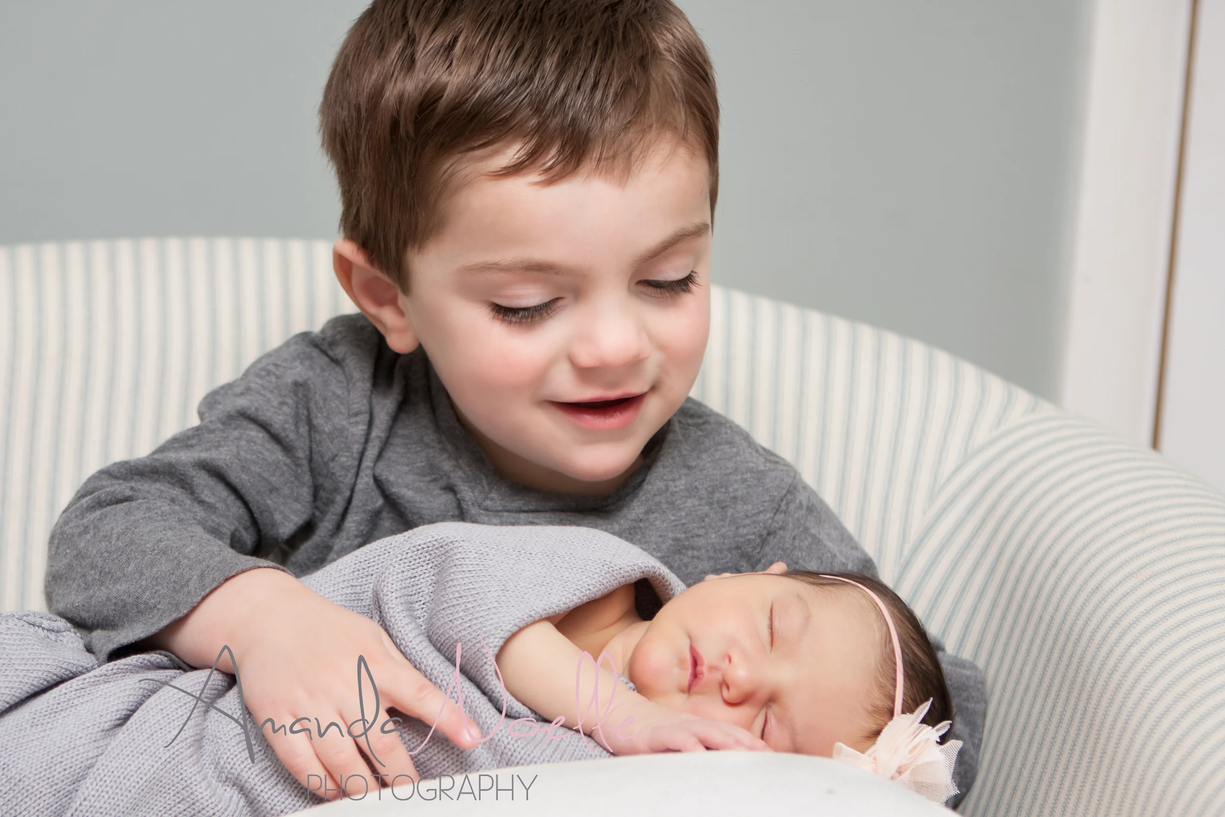 Newborn baby girl, big brother toddler looking at little sister, sibling photography, sitting in chair, newborn on pillow, grey color scheme with pink headband by Westchester, New York Newborn Photographer, Amanda Noelle Photography
