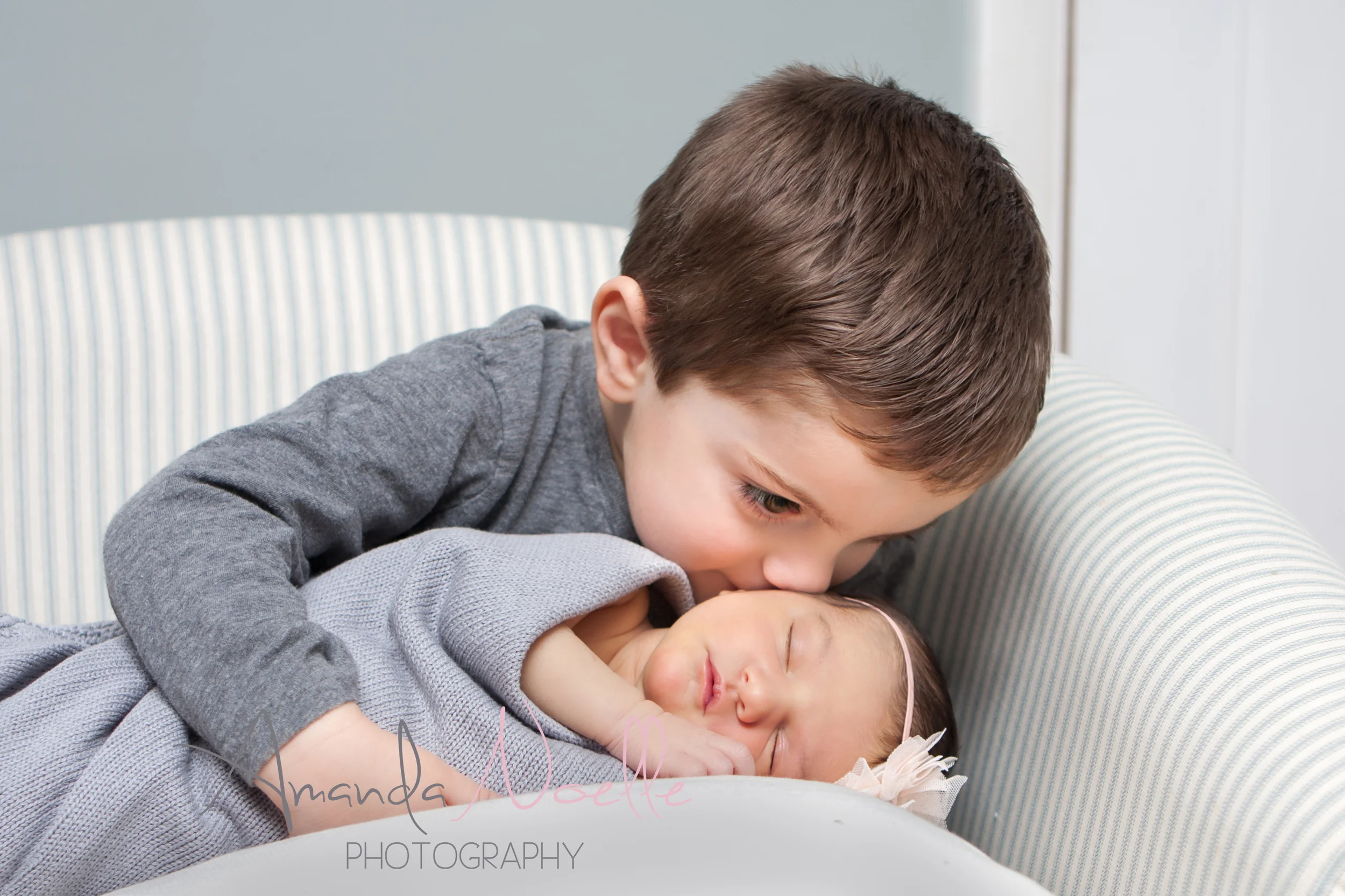 Newborn baby girl, big brother toddler kissing little baby sister, sibling photography, sitting in chair, newborn on pillow, grey color scheme with pink headband by Westchester, New York Newborn Photographer, Amanda Noelle Photography
