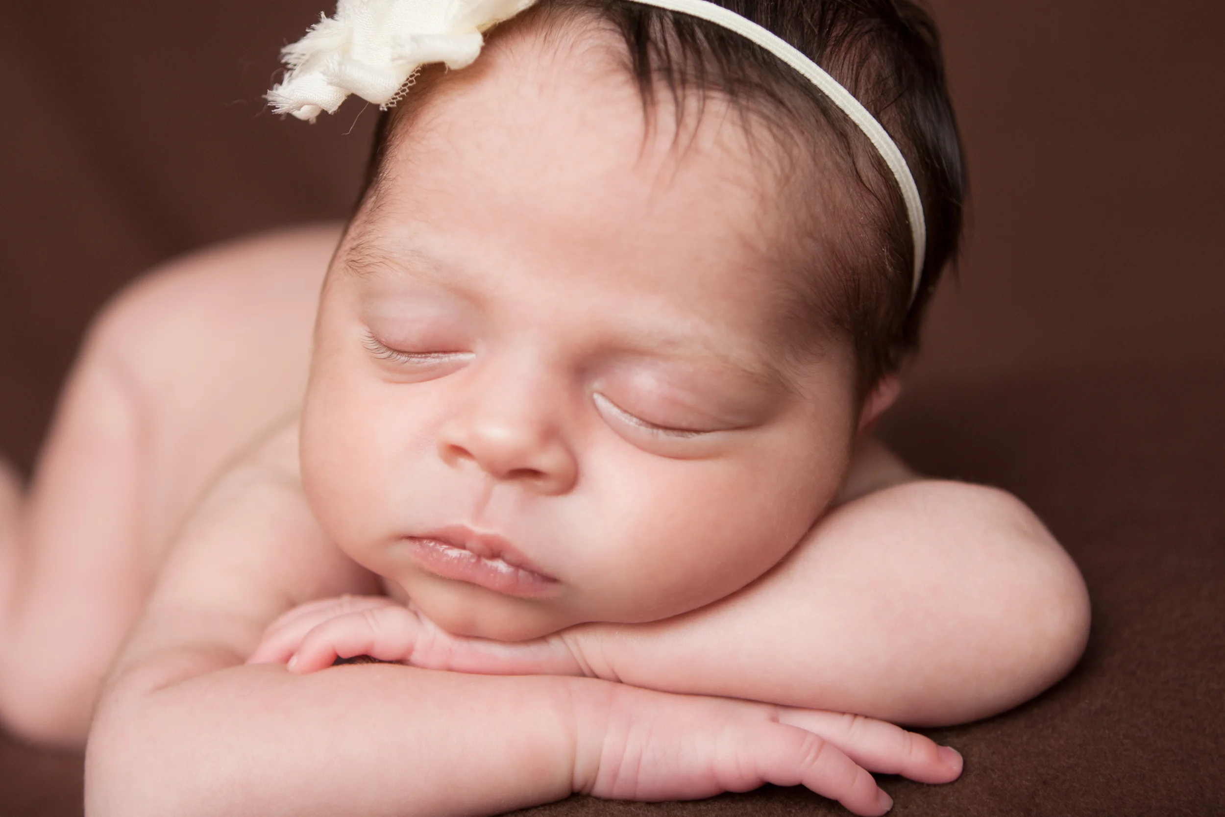 Newborn baby girl, white creme shabby chic flower headband, brown backdrop fabric blanket, head chin on wrists hands, sleeping baby pose on beanbag, Westchester County, New Rochelle, White Plains, Pelham, New York Newborn Photographer, Amanda Noelle Photography