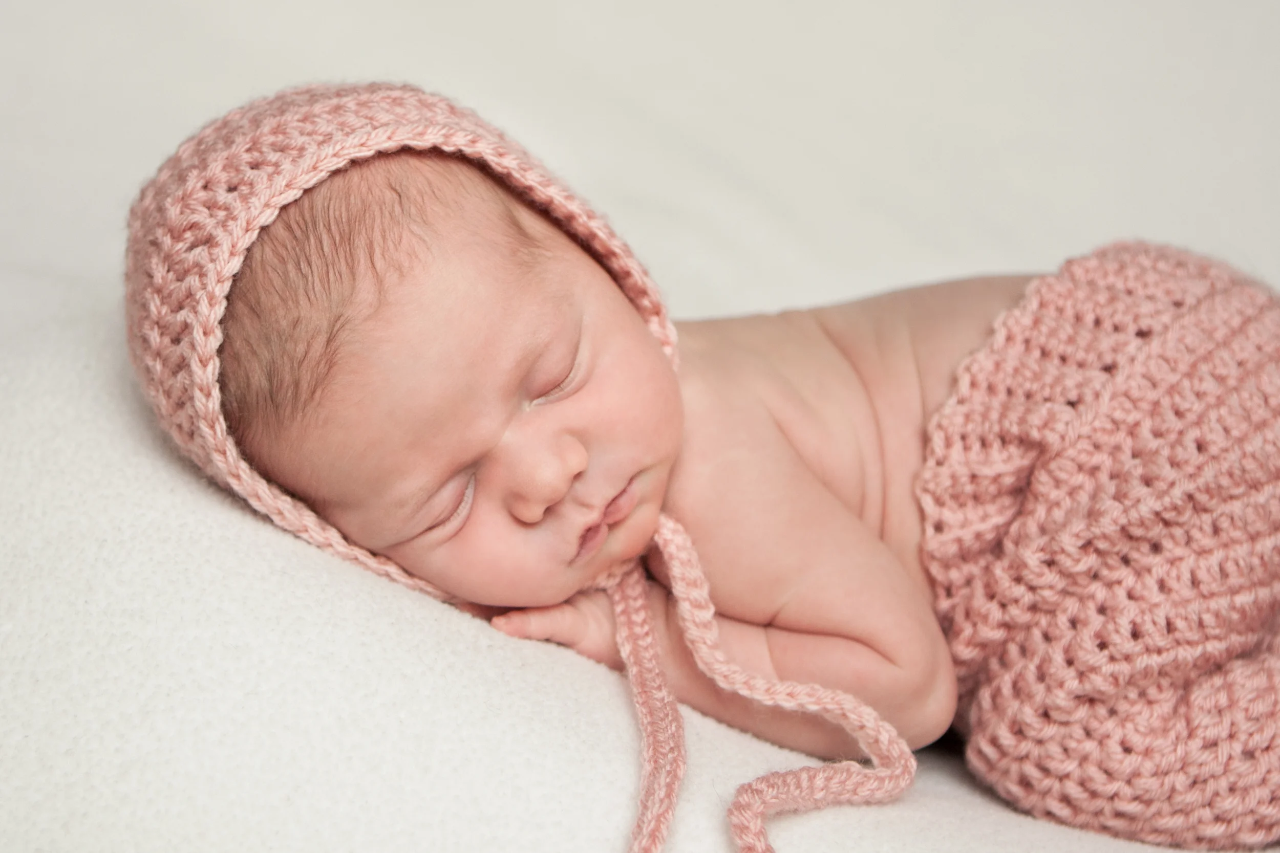Newborn baby girl in crochet pink pants and crochet pink bonnet , bean bag pose, on white fabrick backdrop, Westchester County, New York Newborn Photographer, Amanda Noelle Photography.