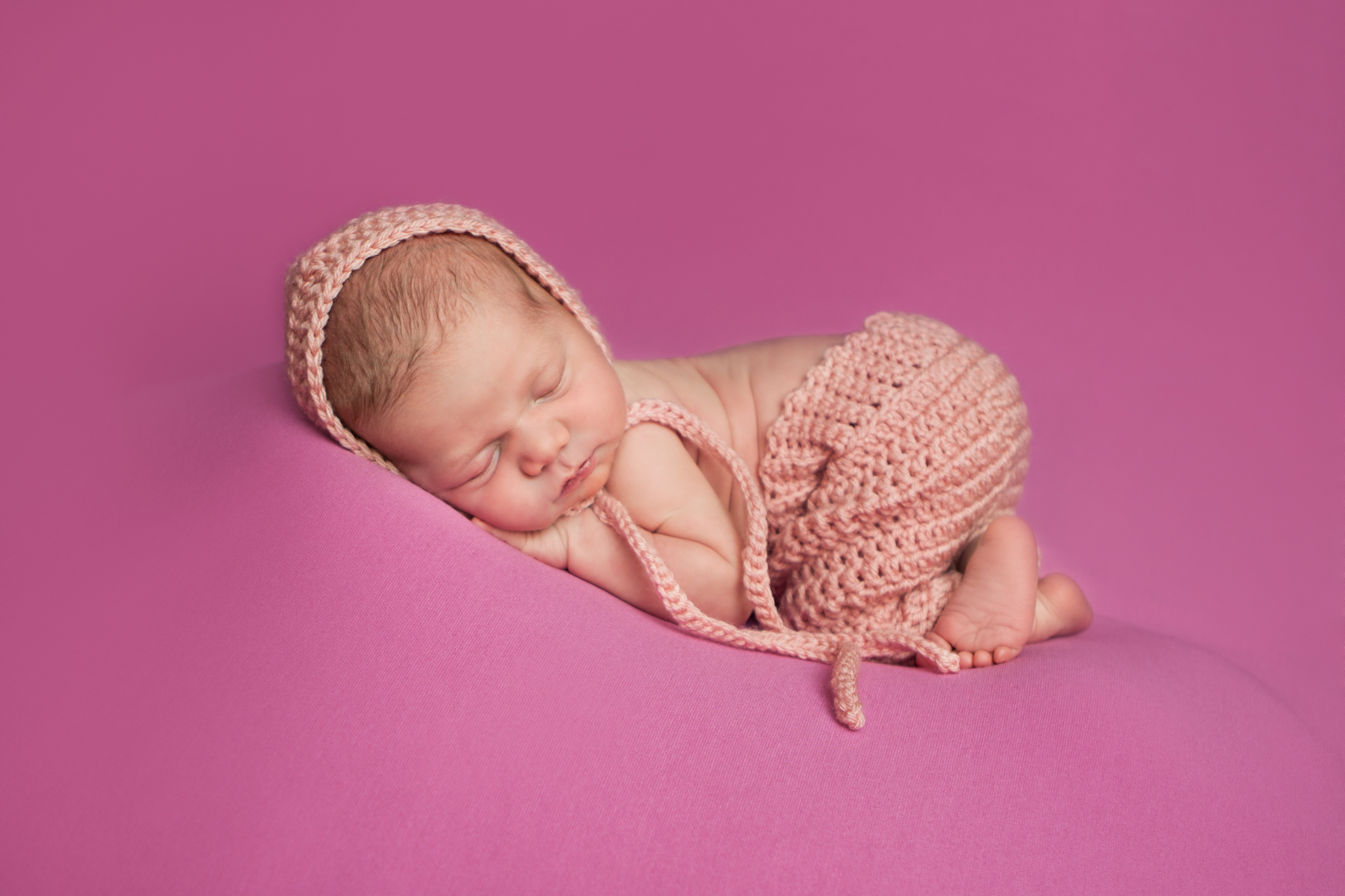 Newborn baby girl in crochet pink pants and crochet pink bonnet , bean bag pose on pink fabric backdrop, Westchester County, New York Newborn Photographer, Amanda Noelle Photography.