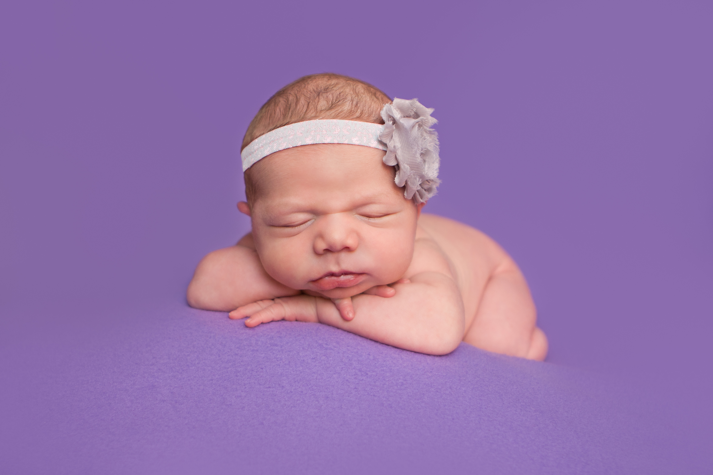 Newborn baby girl with shabby chic headband grey purple, head on wrists chin on hands bean bag pose on purple fabric backdrop, Westchester County, New York Newborn Photographer, Amanda Noelle Photography.