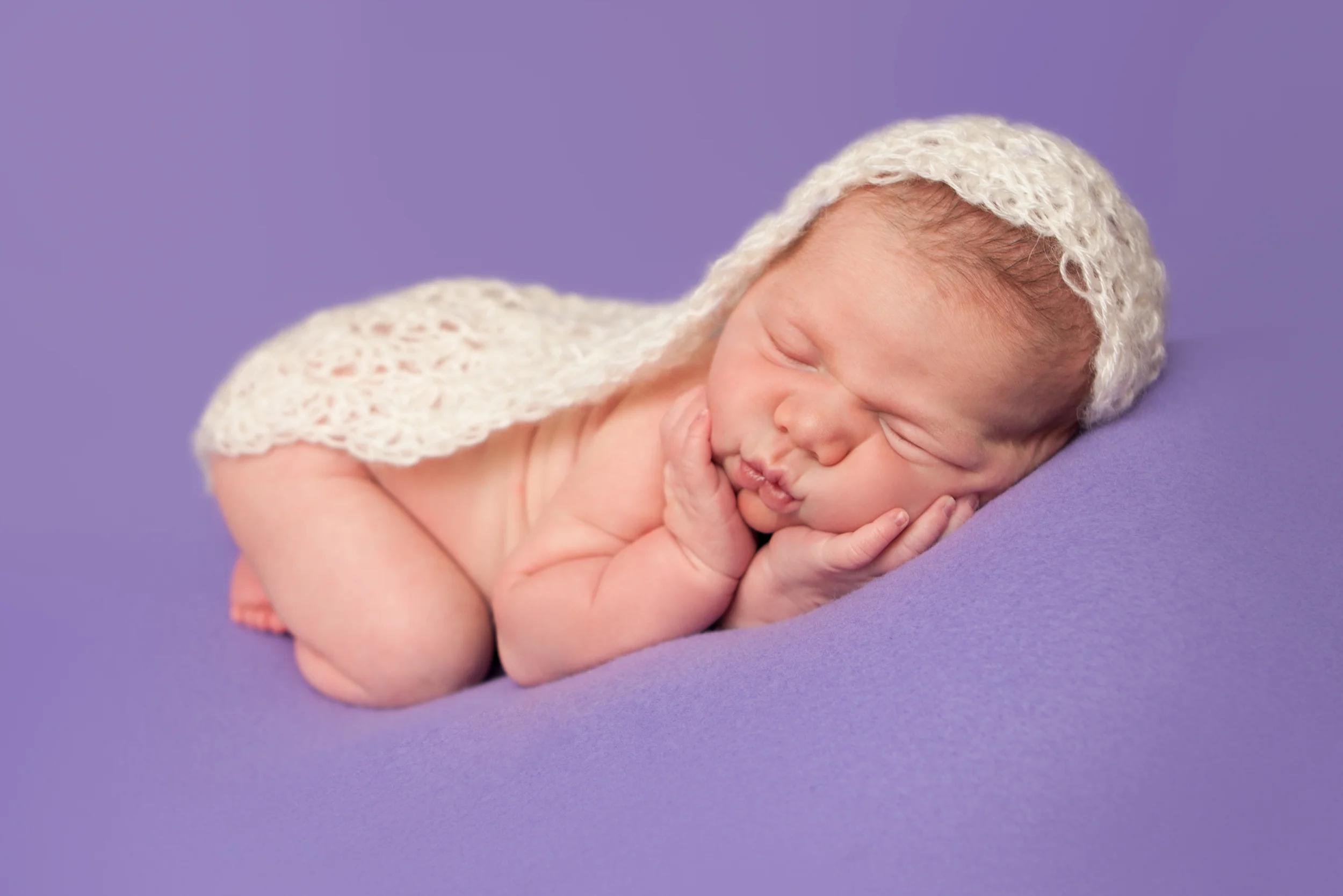 Newborn baby girl with lace crochet wrap, hands on cheeks bean bag pose on purple fabric backdrop, Westchester County, New York Newborn Photographer, Amanda Noelle Photography.