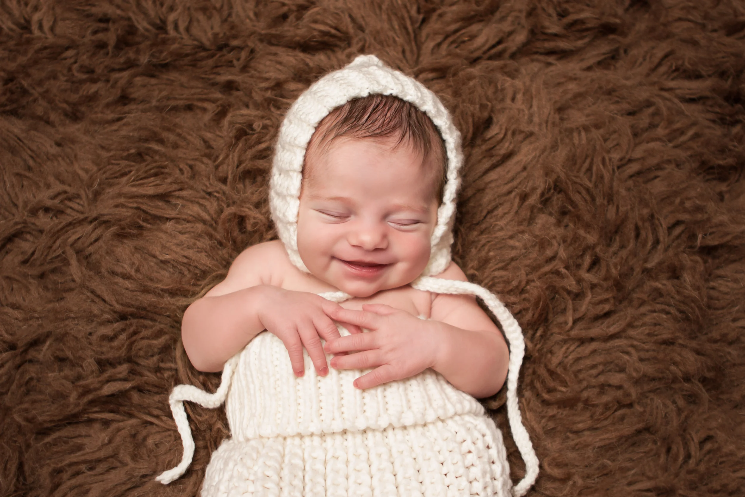 Newborn baby girl smiling , creme white crochet bonnet in, in creme white crochet knit wrap sack on brown flokati rug, Westchester County, New York Newborn Photographer, Amanda Noelle Photography.