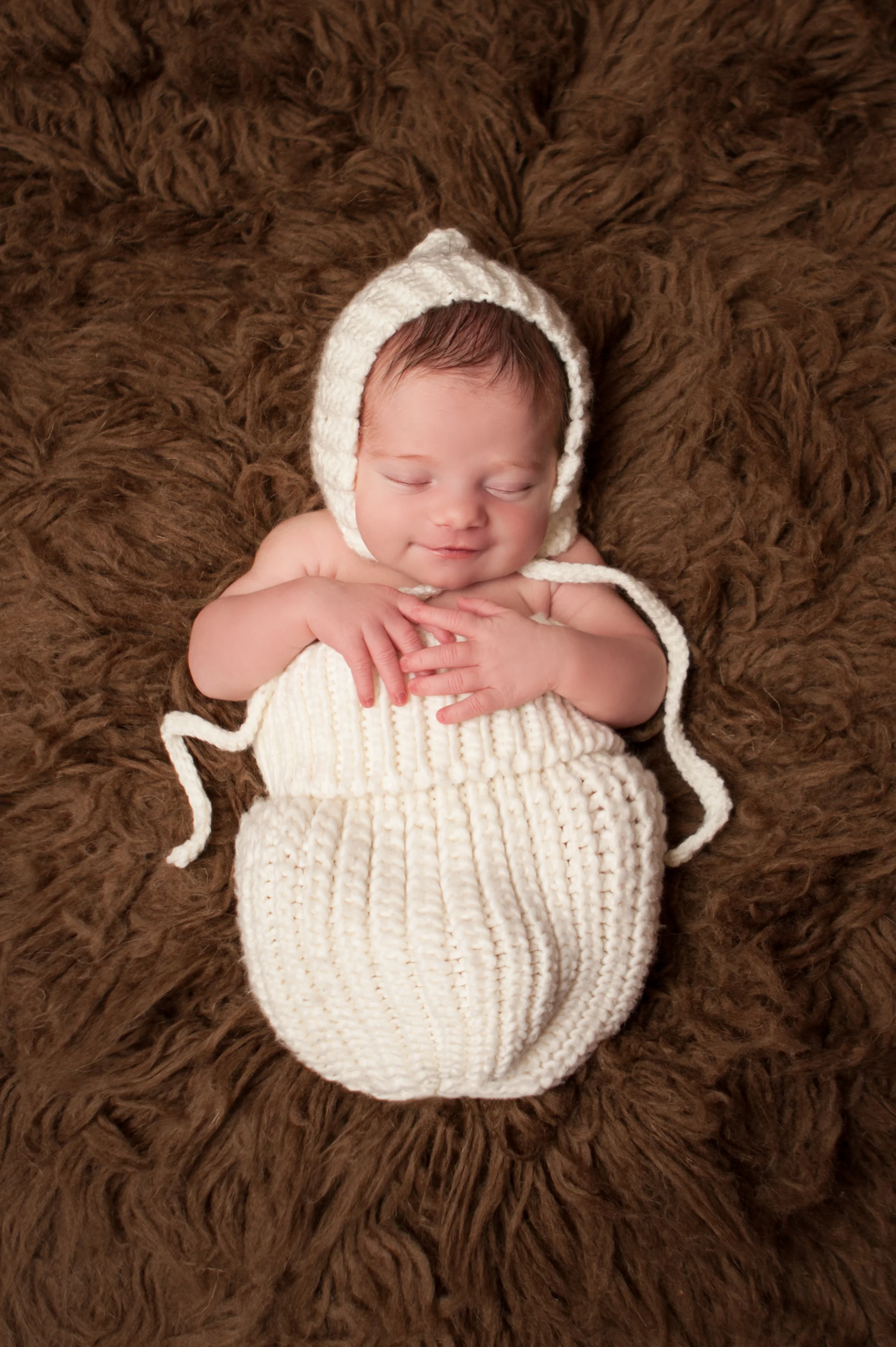 Newborn baby girl smiling , creme white crochet bonnet in, in creme white crochet knit wrap sack on brown flokati rug, Westchester County, New York Newborn Photographer, Amanda Noelle Photography.