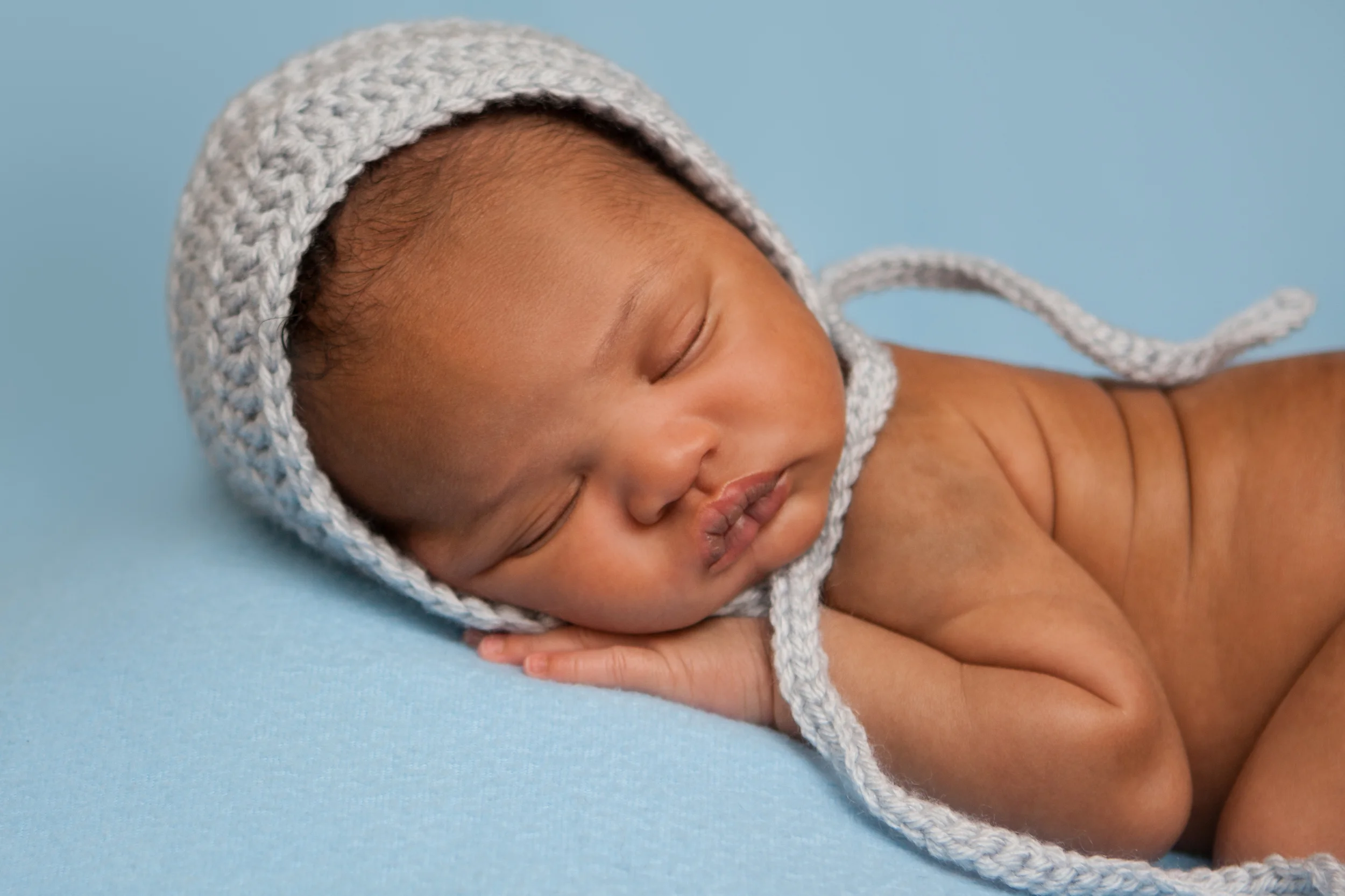 Newborn baby boy j black curly hair wearing blue crochet bonnet, african american, tushy up sleeping bean bag pose, blue fabric blanket backdrop, Westchester County, New York Newborn Photographer, Amanda Noelle Photography.