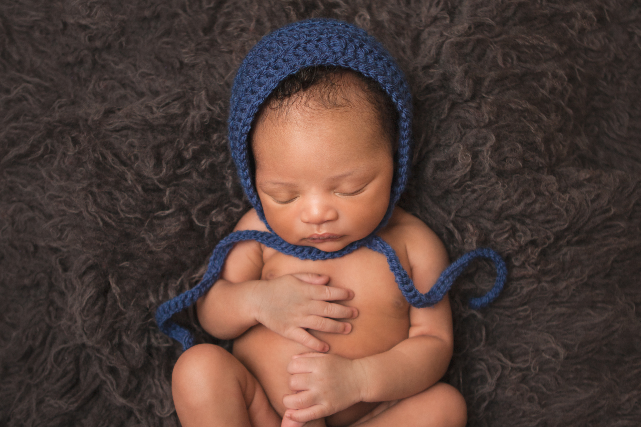 Newborn baby boy j black curly hair wearing dark blue crochet bonnet, african american, sleeping pose holding foot on grey gray blue flokati rug , Westchester County, New York Newborn Photographer, Amanda Noelle Photography.