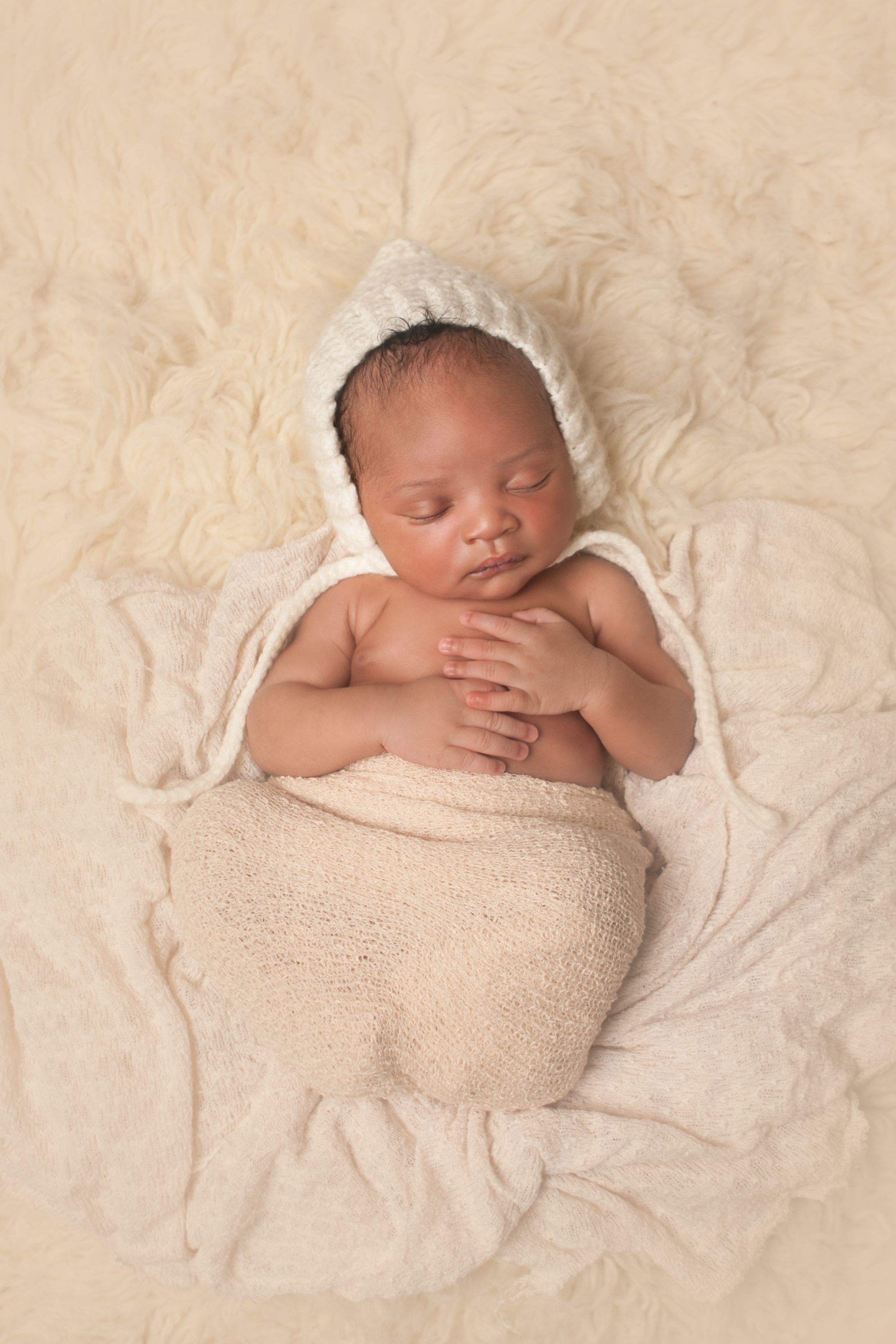 Newborn baby boy j black curly hair, african american, wearing cream white crochet bonnet, wrapped in cream stretch knit wrap, sleeping on cream flokati rug, Westchester County, New York Newborn Photographer, Amanda Noelle Photography.