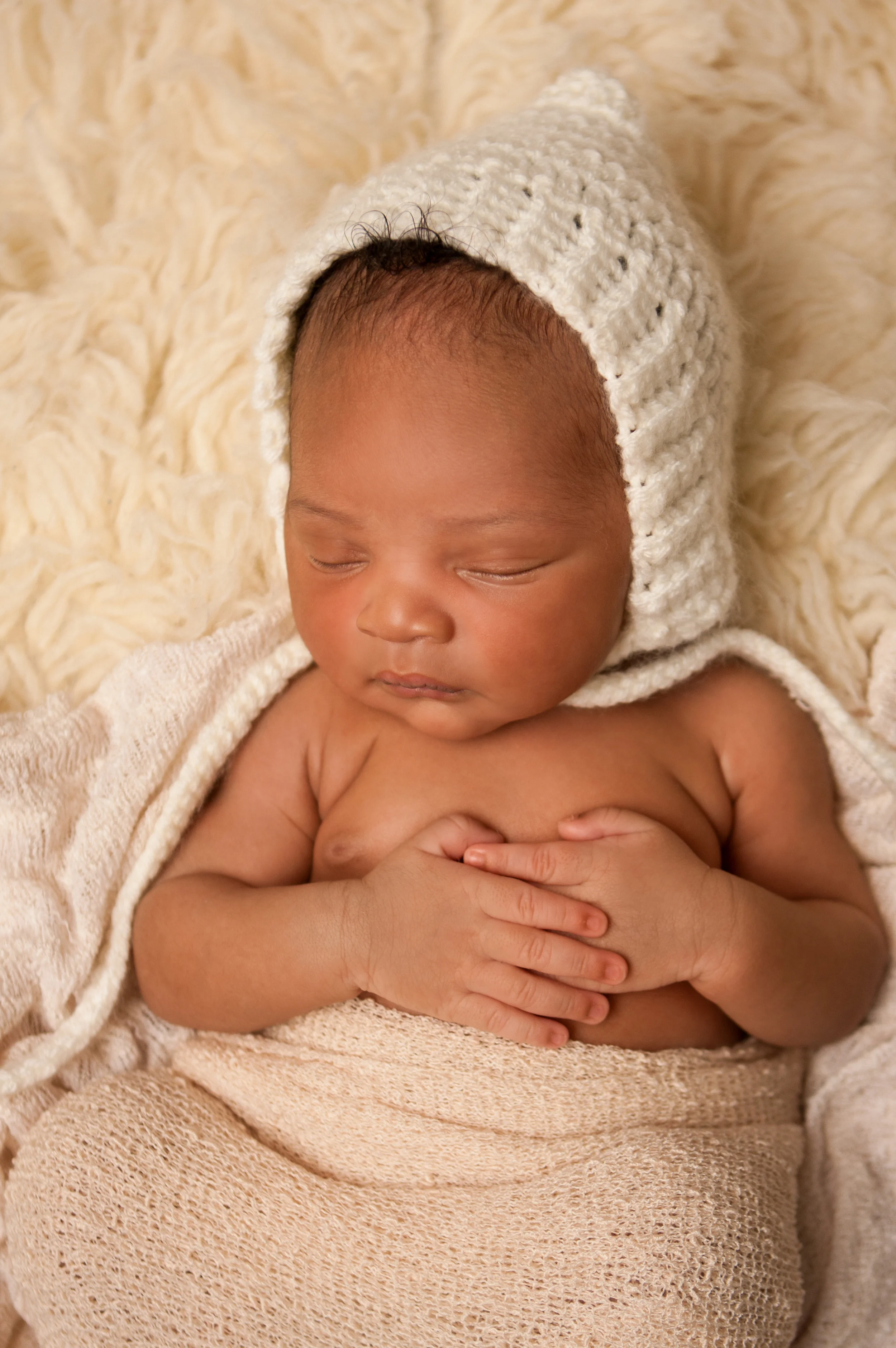 Newborn baby boy j black curly hair, african american, wearing cream white crochet bonnet, wrapped in cream stretch knit wrap, sleeping on cream flokati rug, Westchester County, New York Newborn Photographer, Amanda Noelle Photography.