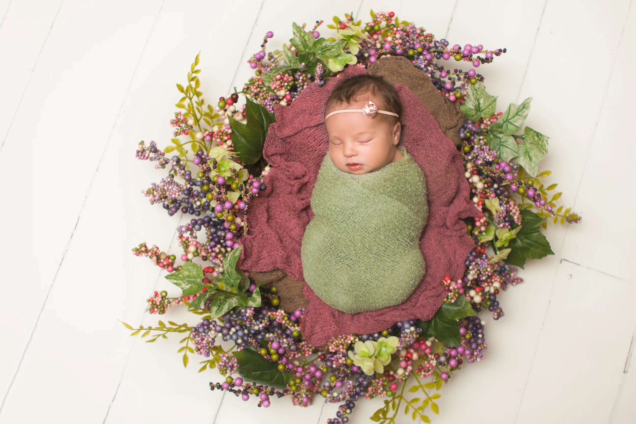 Newborn baby girl sleeping spring photos, wrapped in green knit stretch wrap, spring wreath, white wood floor backdrop.