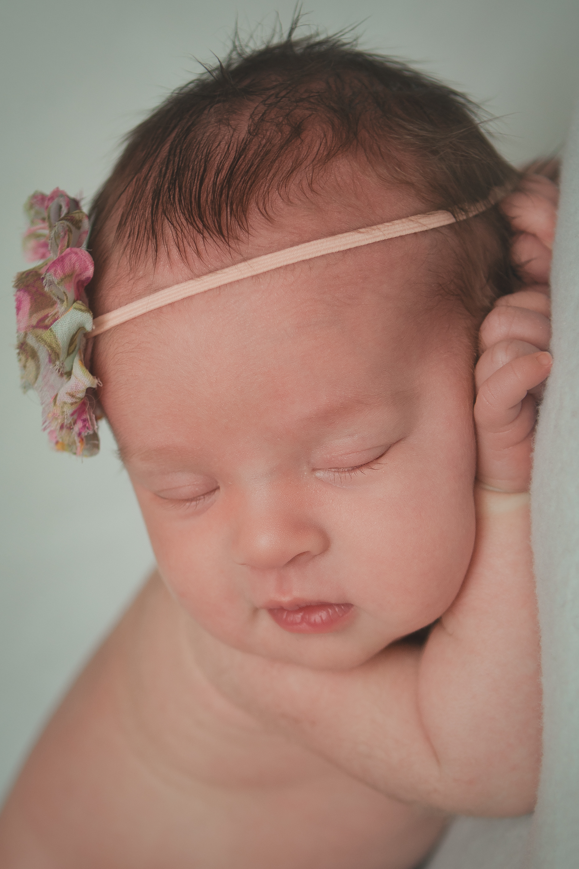 Newborn baby girl sleeping pose, rose pink flower headband, green blue blanket fabric backdrop, head chin on hands bean bag pose.