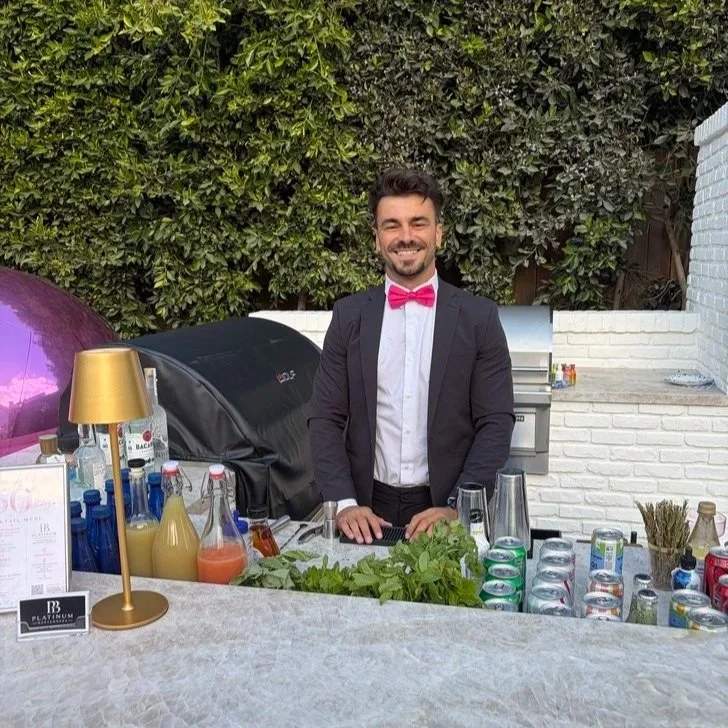 A smiling man in a black suit with a pink bow tie stands behind a bar area outdoors. The bar has various bottles, cans, and mixers, with some greenery and a grill in the background.