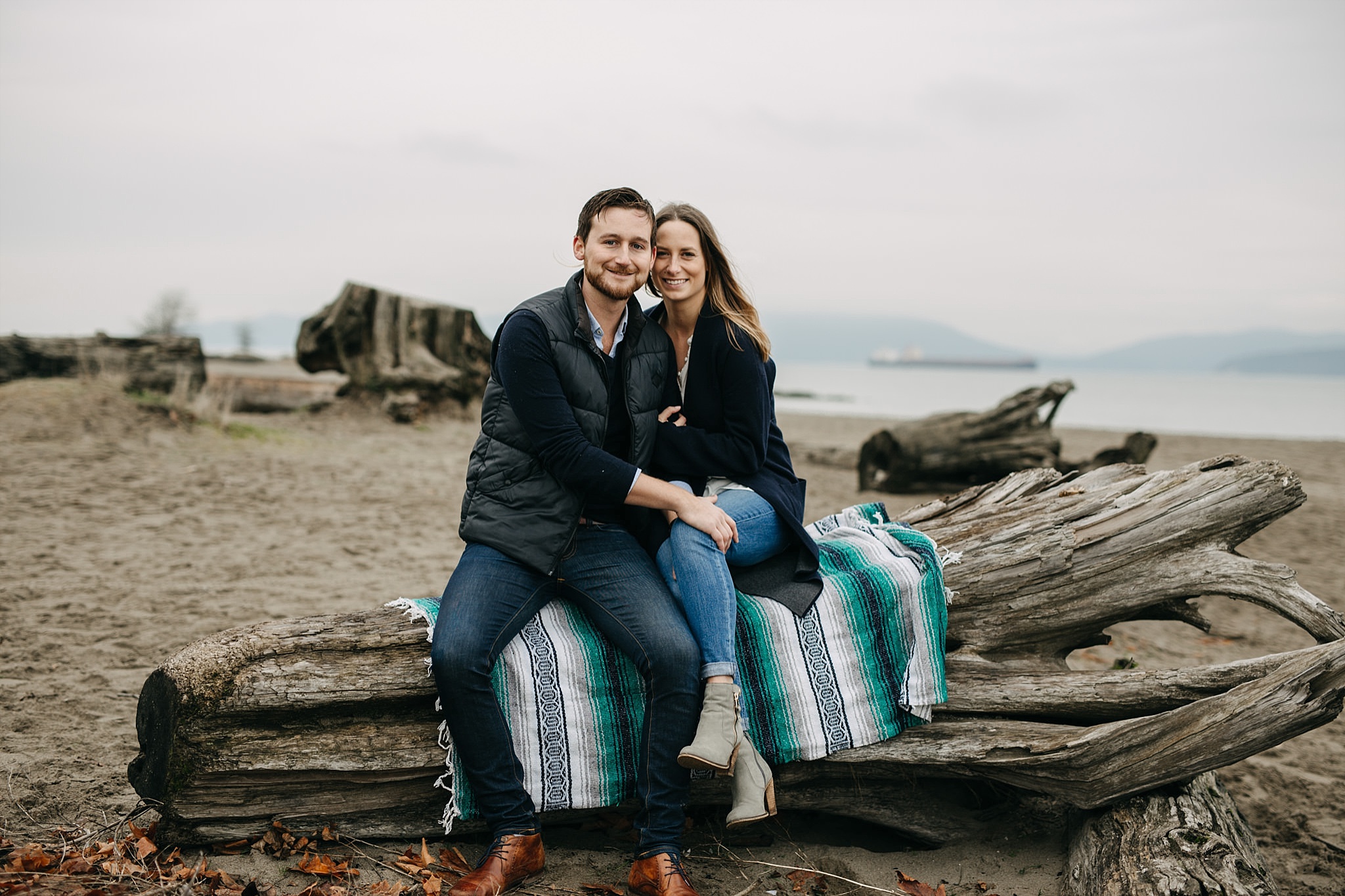 couple smiling sitting on blanket on log at spanish banks beach engagement