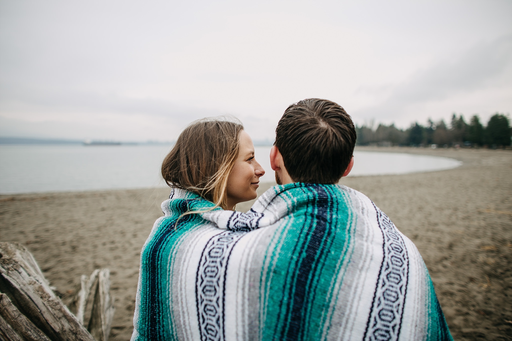 couple wrapped in blanket sitting on log at spanish banks beach engagement