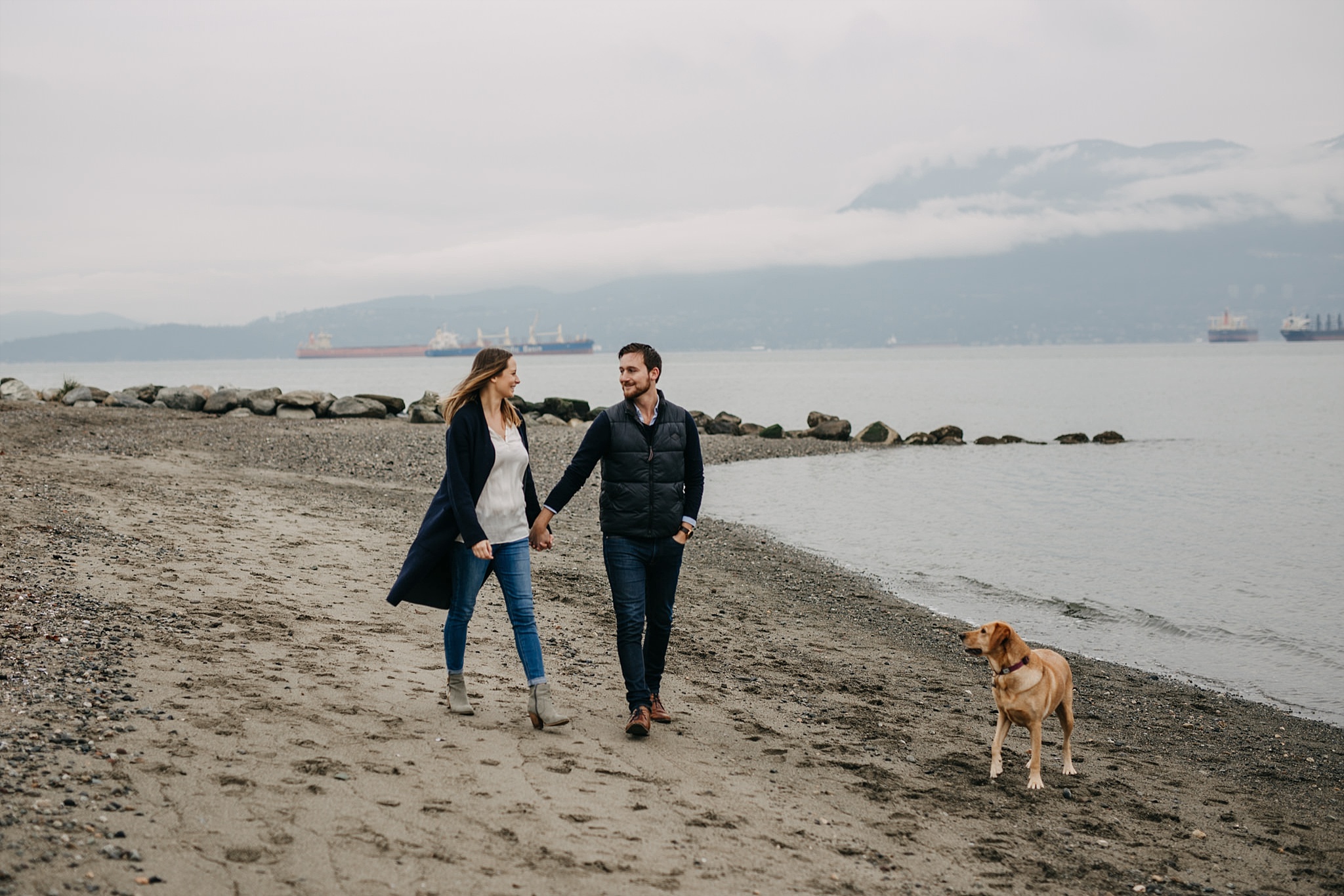 couple holding hands walking on beach with puppy spanish banks engagement