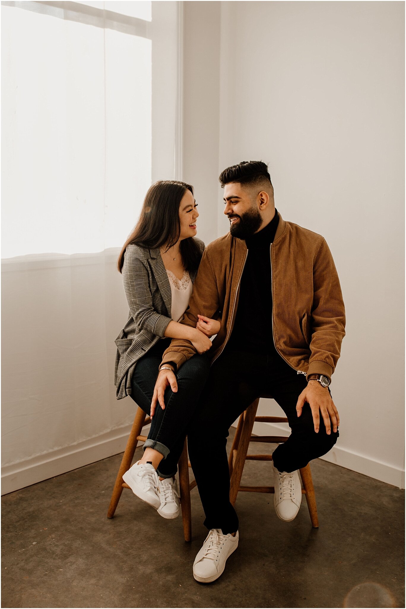 interracial couple sitting on wooden stools smiling portrait in natural light studio in vancouver