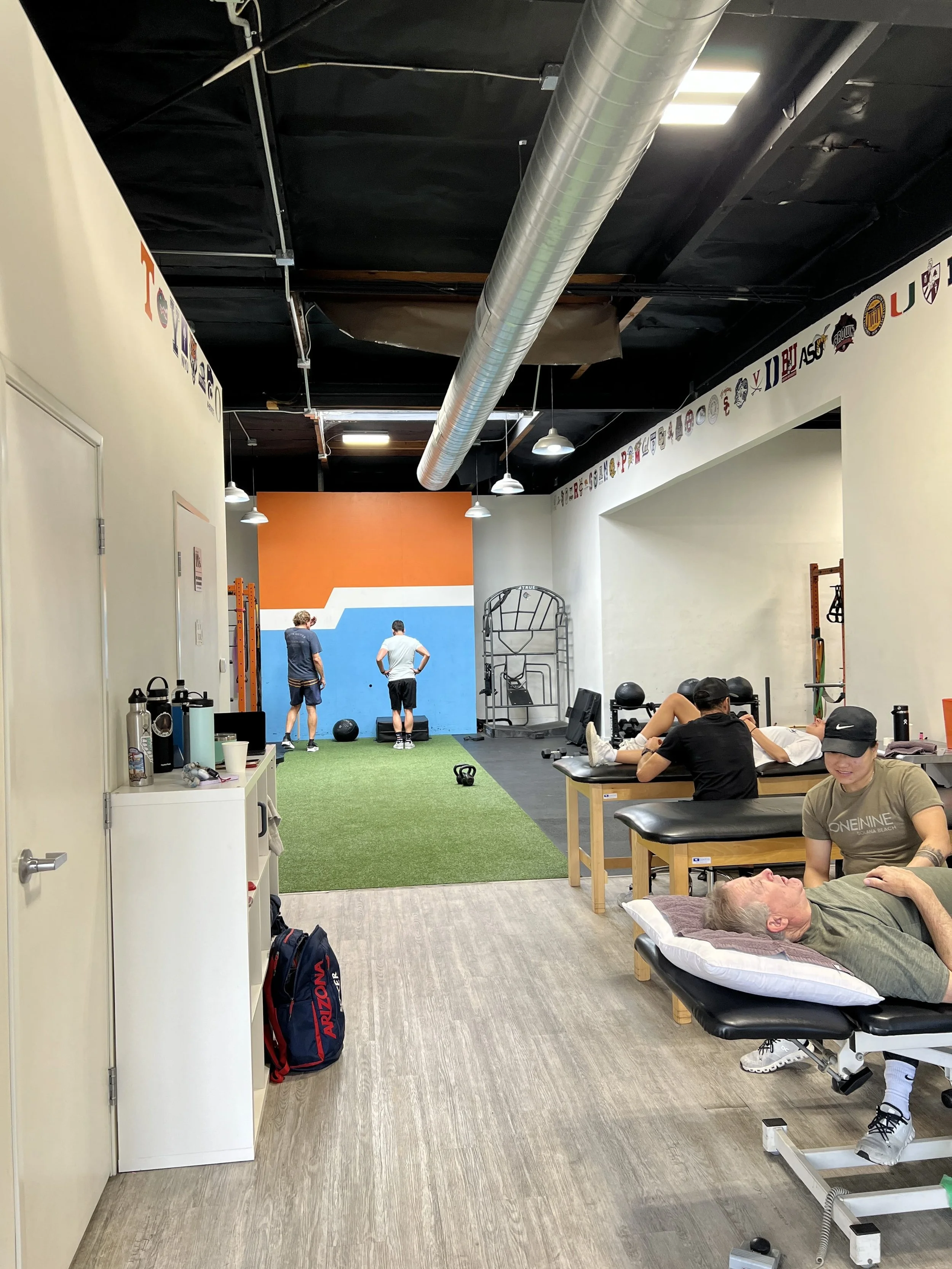 A gym interior with people working out and receiving physical therapy. Some individuals are performing exercises on the turf, while one person is lying on a treatment table being attended to by a therapist. The space has a colorful wall at the back, gym equipment, and a shelf with water bottles and casual items.