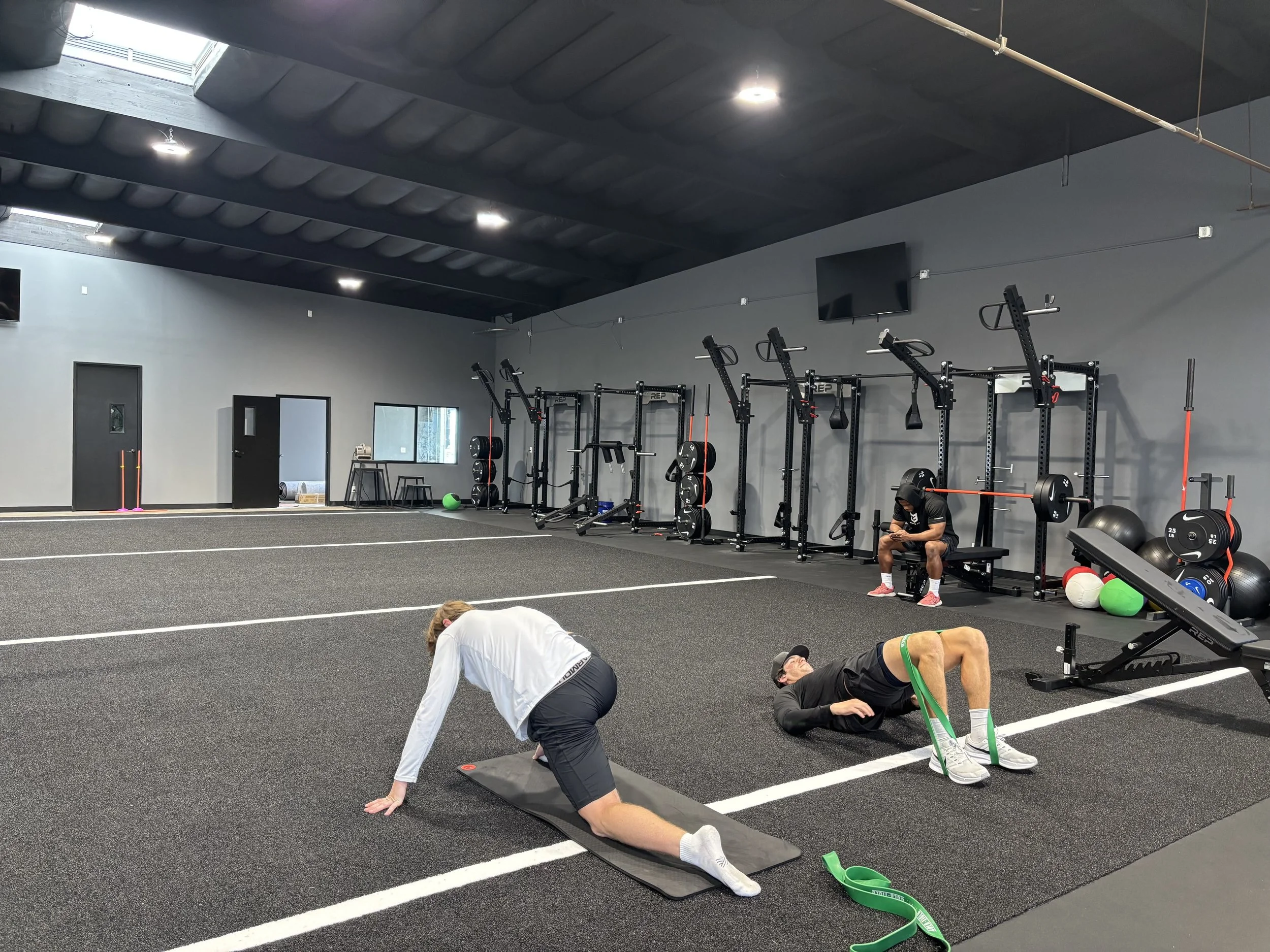 Two people working out on a black rubber gym floor with white lines, a woman performing a stretch and a man doing glute bridge exercises with resistance bands. In the background, there are weight racks, workout equipment, and a person sitting on a bench.
