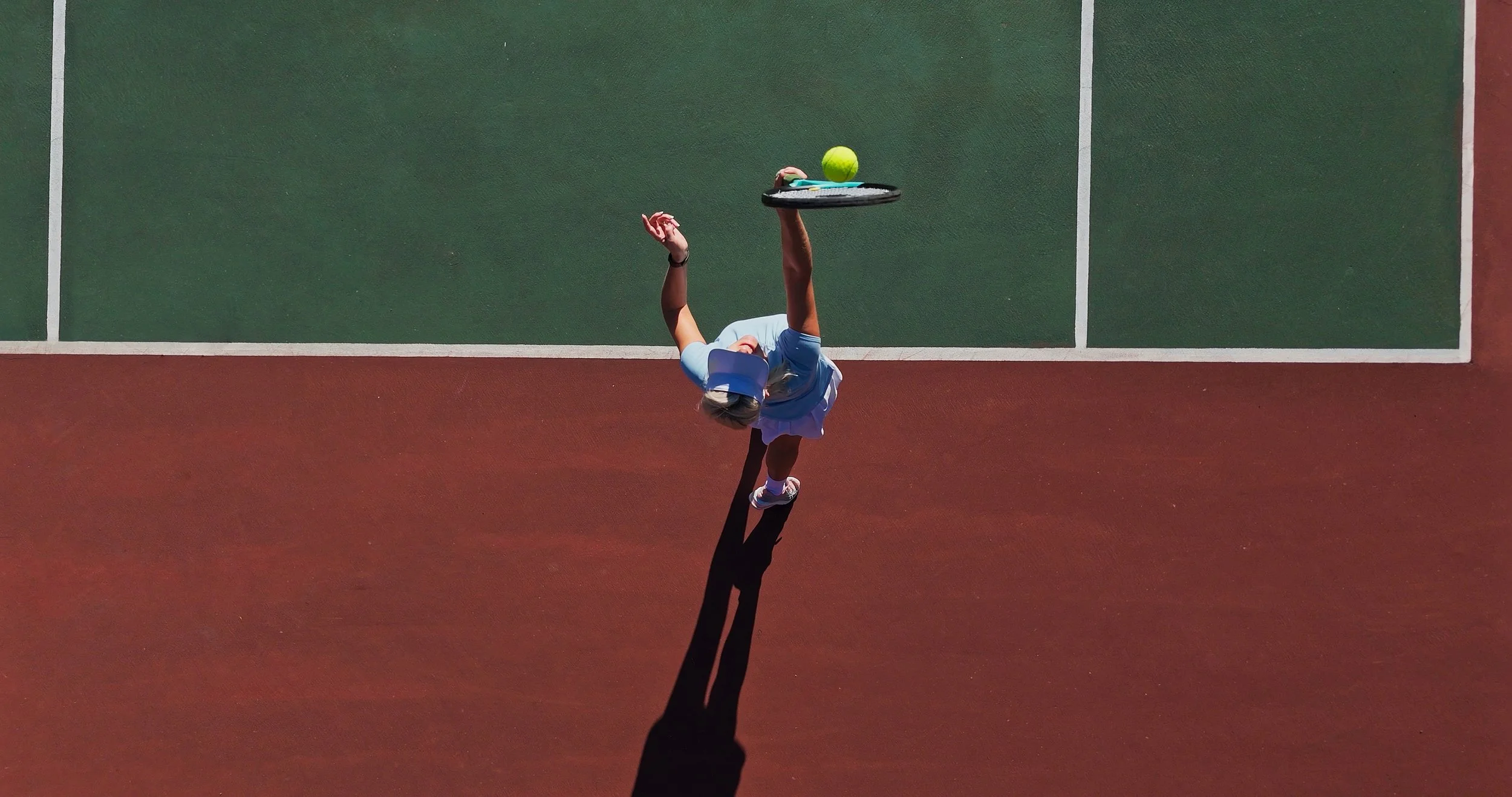 An overhead shot of a tennis player in a light blue shirt and white skirt serving on a tennis court, casting a long shadow on the red surface.