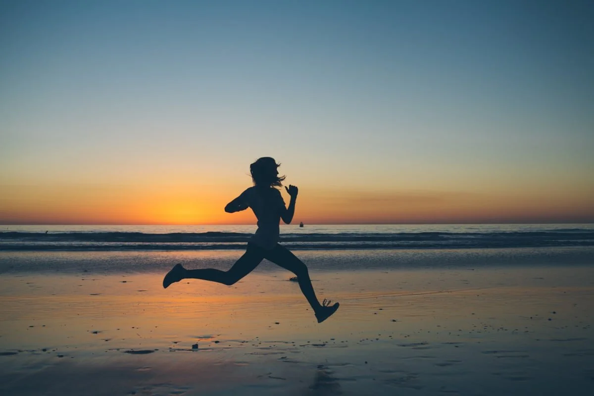 A person running on the beach during sunset, with the ocean and a boat in the background.