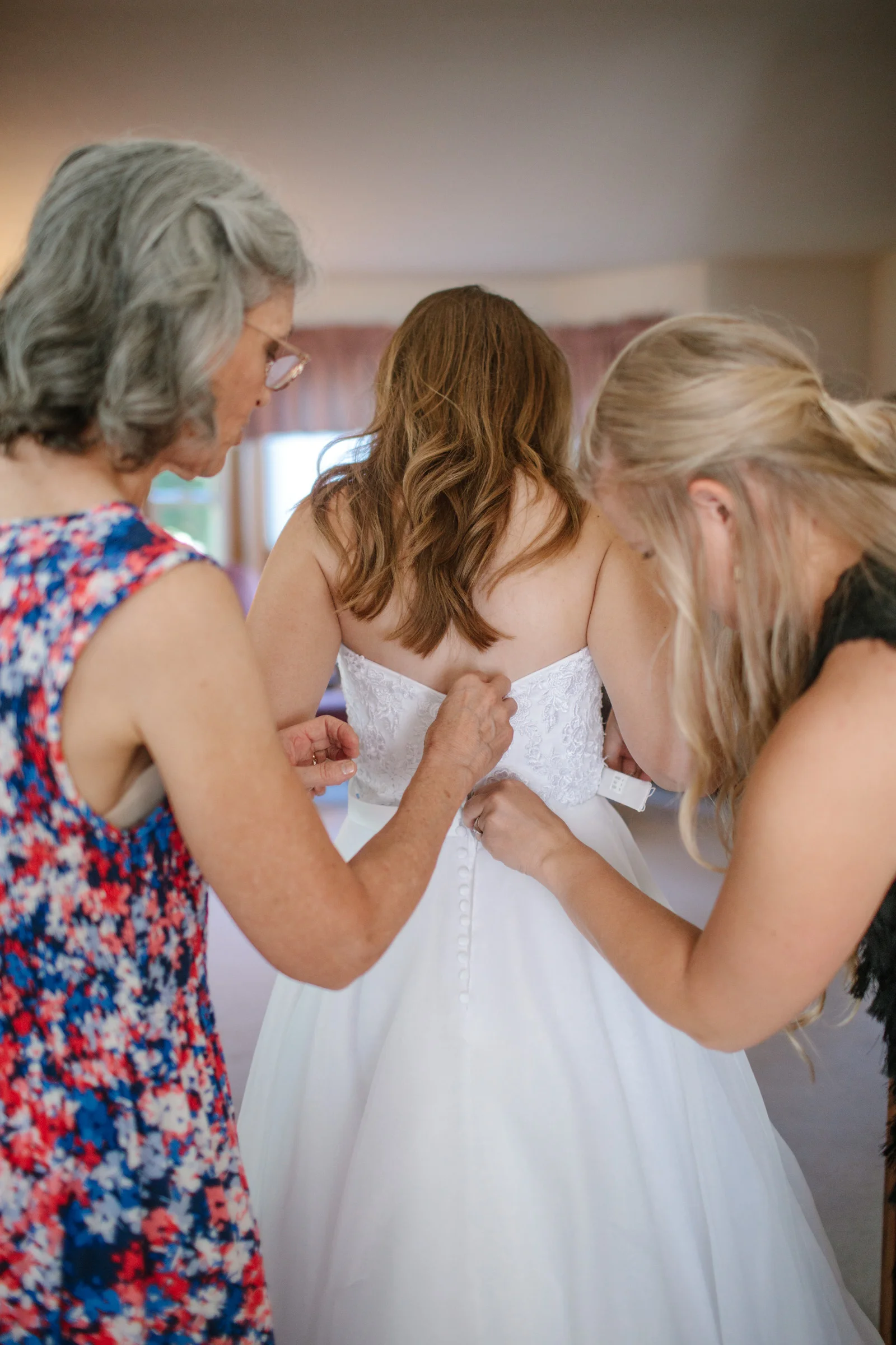 mom and matron of honor helping bride libby put her dress on