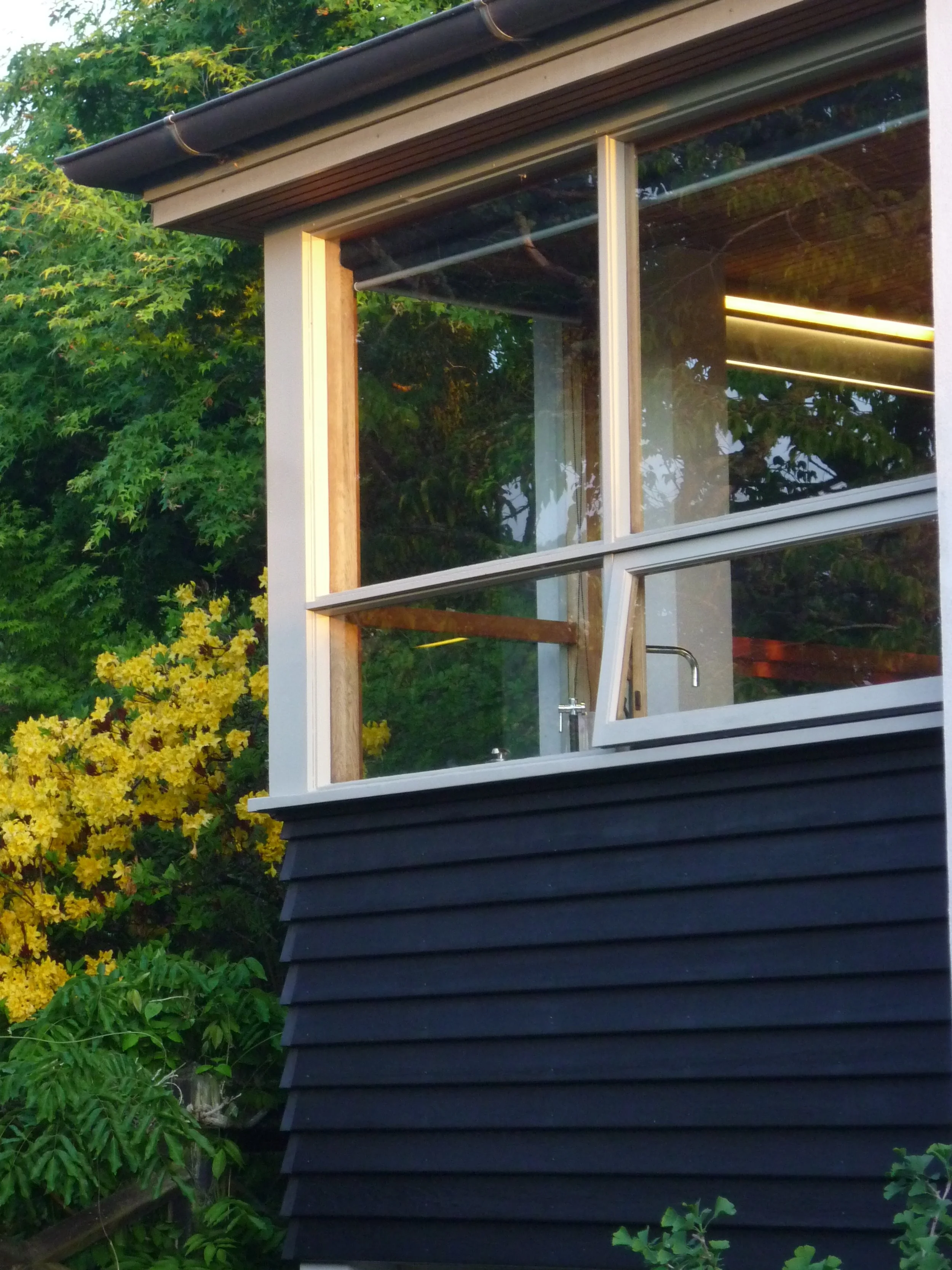 Close-up of a modern house corner with open sash window, black horizontal siding, and a background of green trees and yellow flowers.