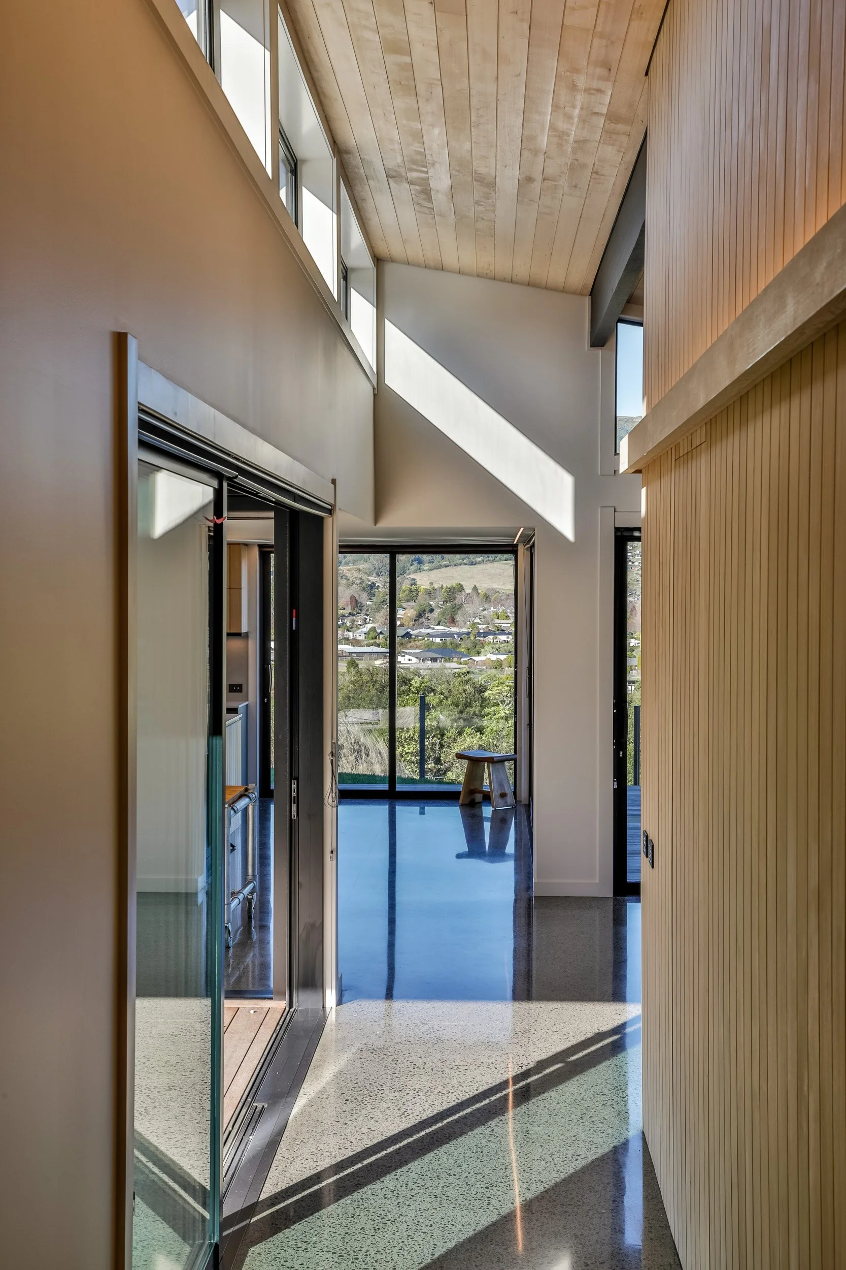 Interior view of a modern house with high ceilings, large windows, and a sliding glass door leading outside. Sunlight casts shadows on the polished floor. Kinloch Architects. Taupo Architects. Home Design. Interior Design. New Zealand Architects. 