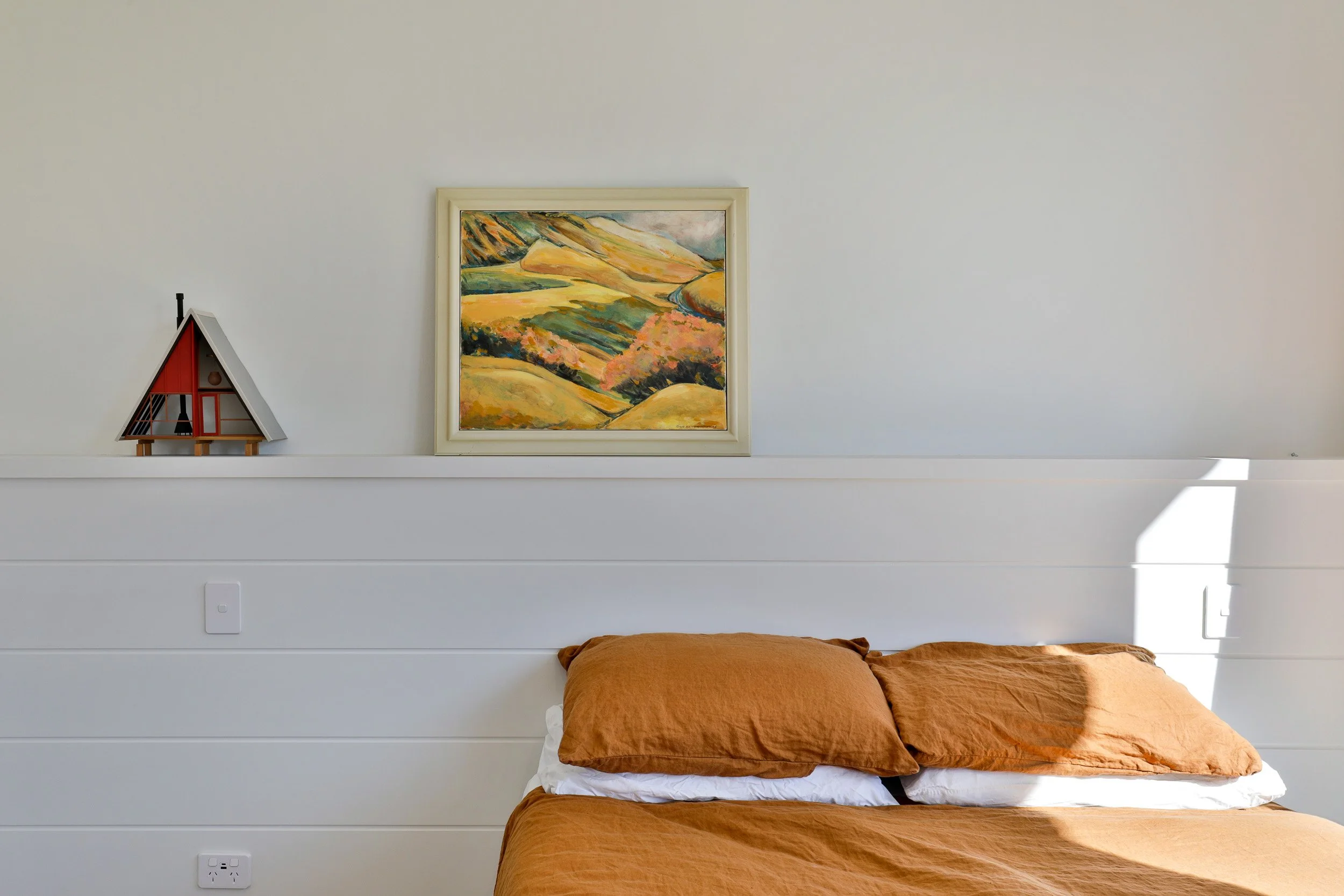A bedroom with a beige bed with orange pillows and a white bedsheet, white shiplap walls, a landscape painting with green and yellow hills, a small decorative house on a shelf, and sunlight casting a shadow on the wall.