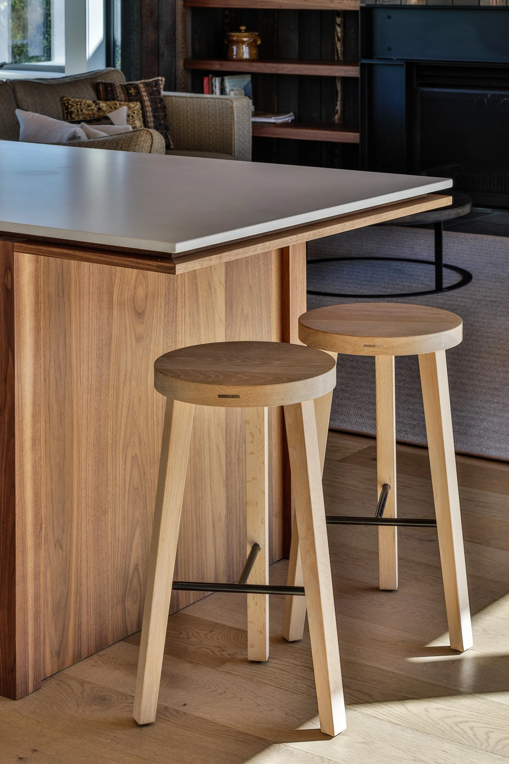 Interior of a room with a wooden kitchen island and two wooden bar stools. In the background, there is a cozy seating area with a sofa, pillows, and a bookshelf.