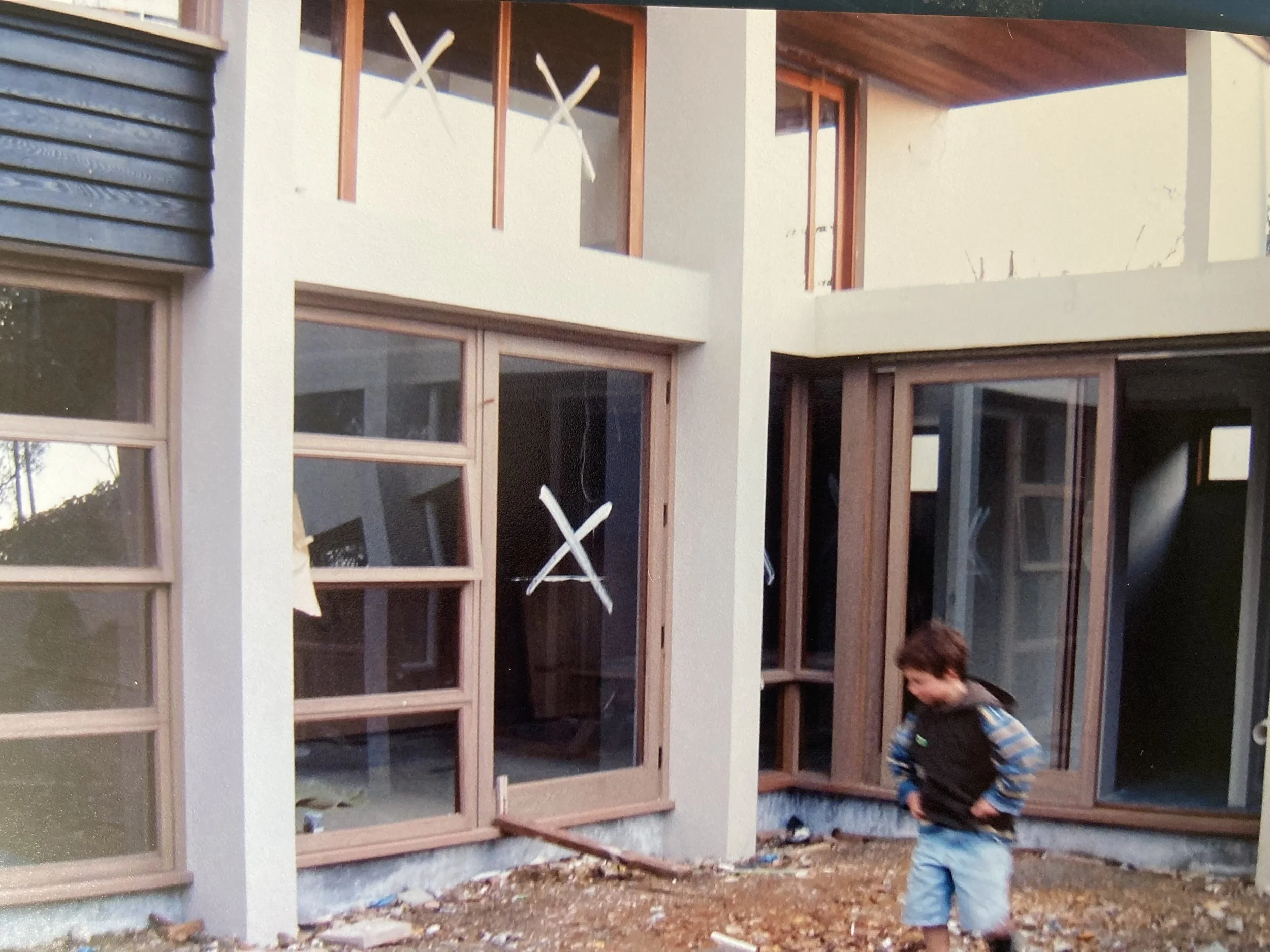 A young boy standing outside a modern house under construction, with large glass windows and a second-floor balcony, surrounded by dirt and construction debris.