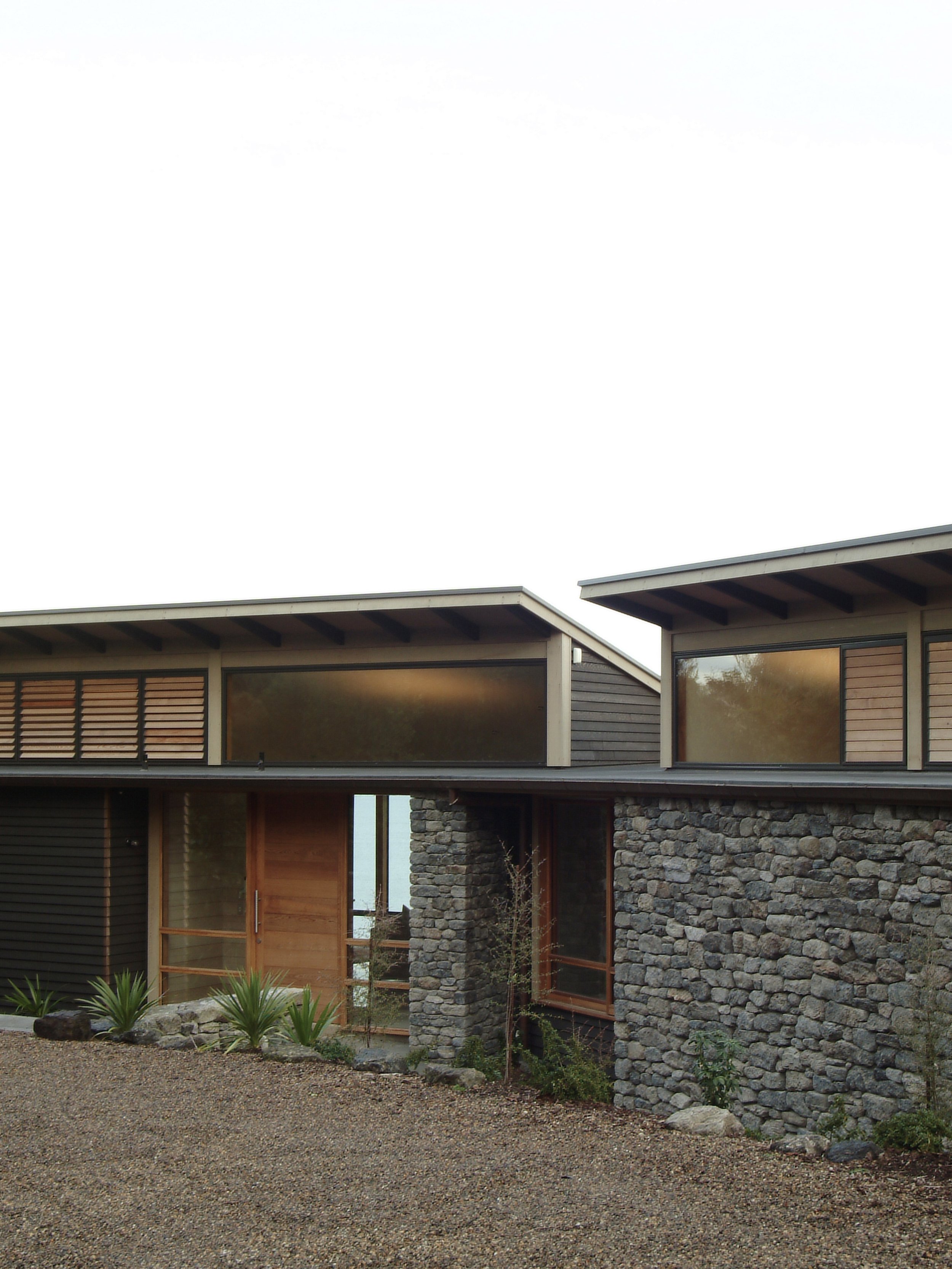 Modern house with stone and wood exterior, large windows, and a gravel front yard with plants.