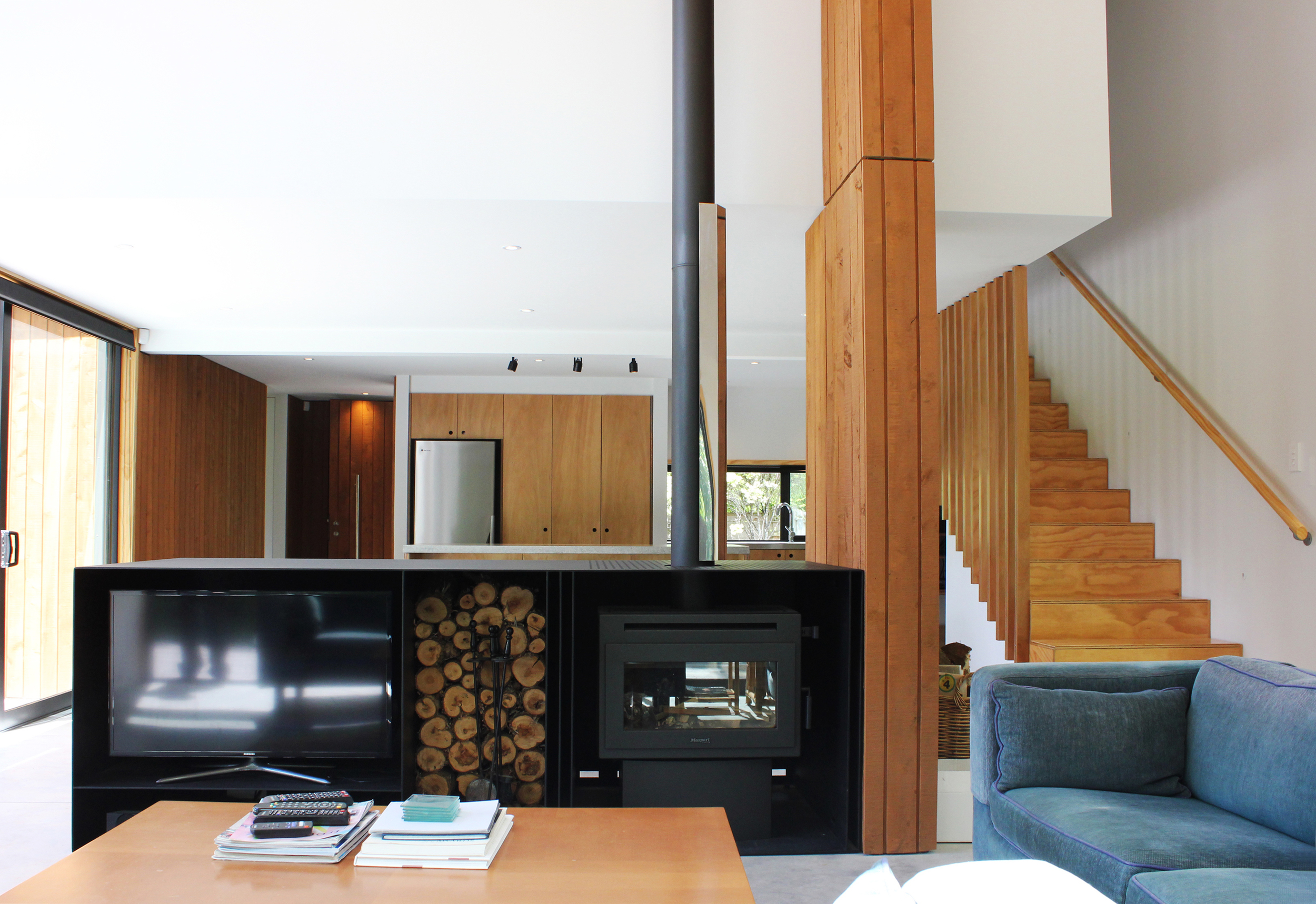 Living room with a wood-burning stove, wood logs, and modern furniture, including a blue sofa and wooden accents. Bach Design New Zealand. Bach Architect. Holiday Home Architect. Rural Architect. 