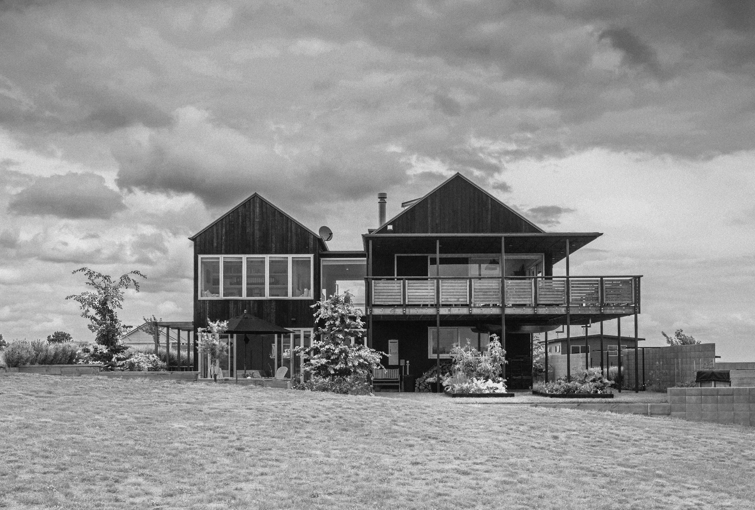 A modern two-story house with a dark exterior, large windows, and a spacious balcony, set in a landscape with trees and a grassy yard under a cloudy sky.