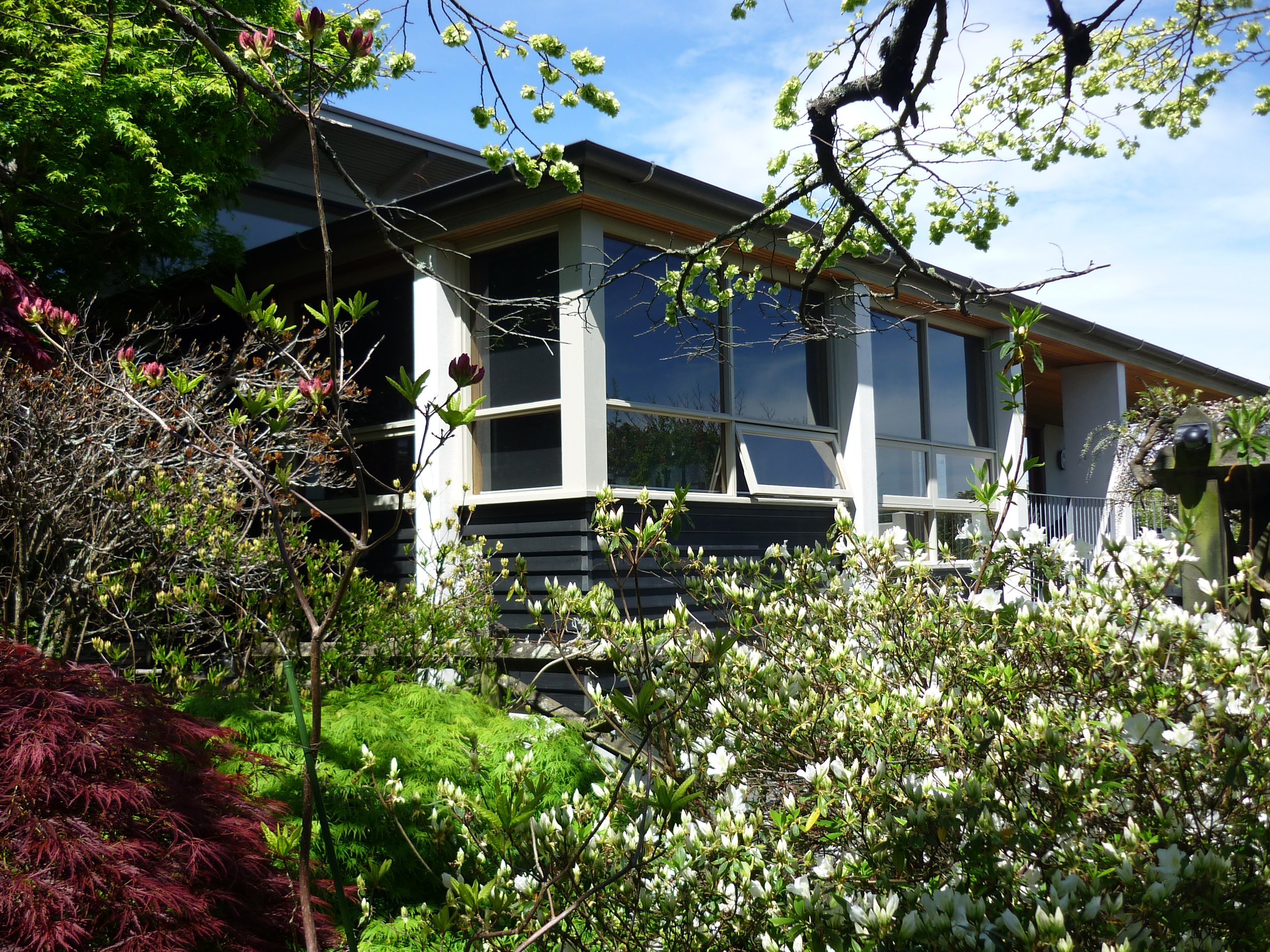 A modern house with large windows surrounded by blooming plants and trees during daytime.