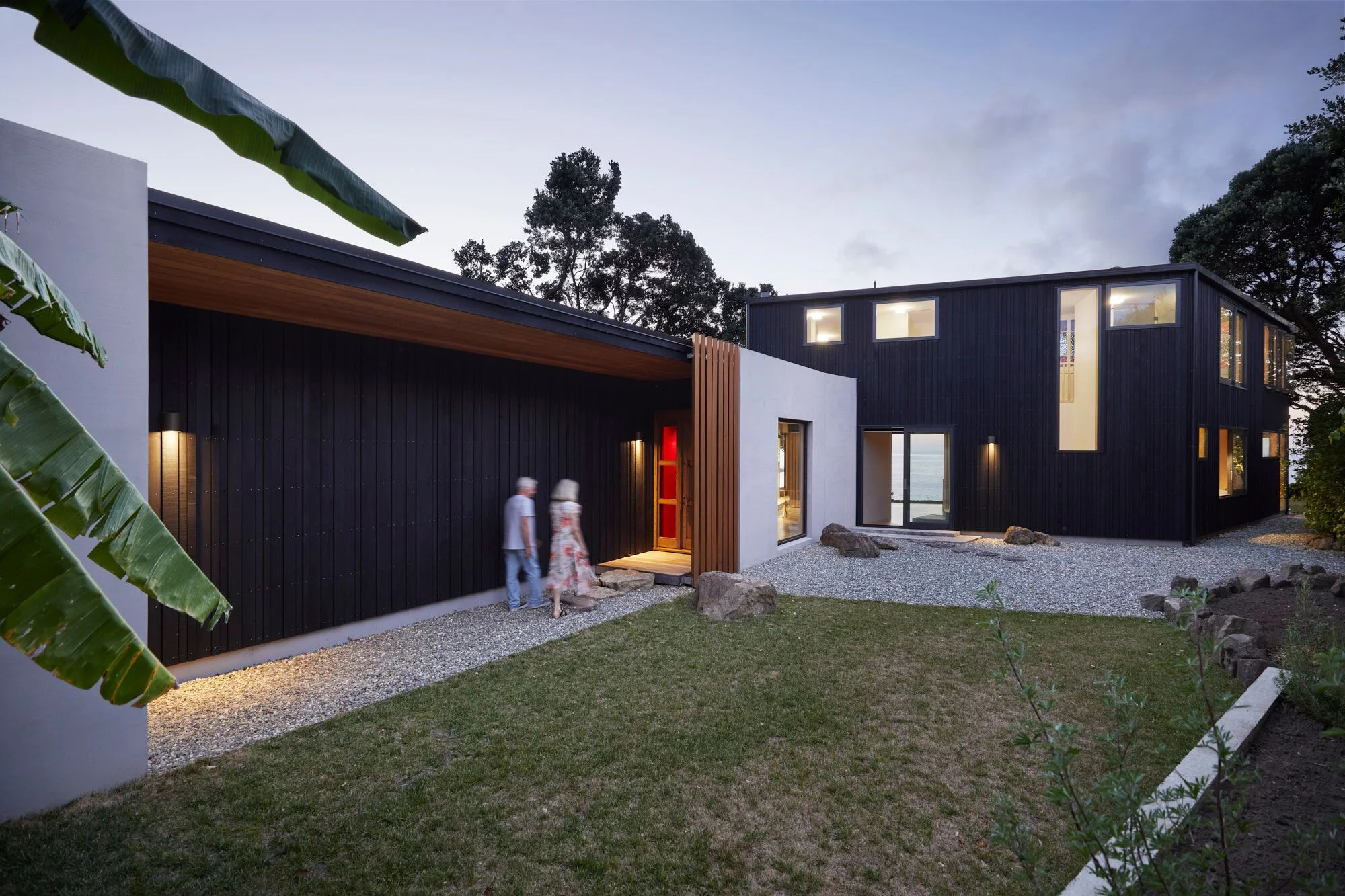 Modern black and white house with a gravel pathway, two people walking, greenery, trees, and a cloudy sky at dusk.